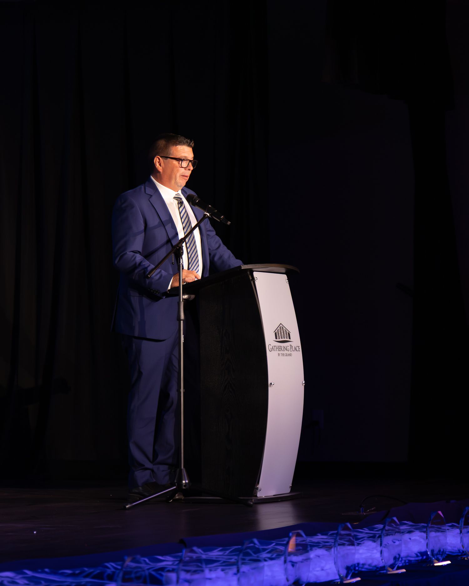 A speaker presents at a podium on stage with blue lighting effects.