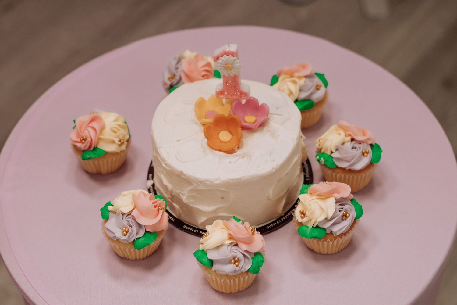 White birthday cake surrounded by floral cupcakes arranged on a pink serving plate.