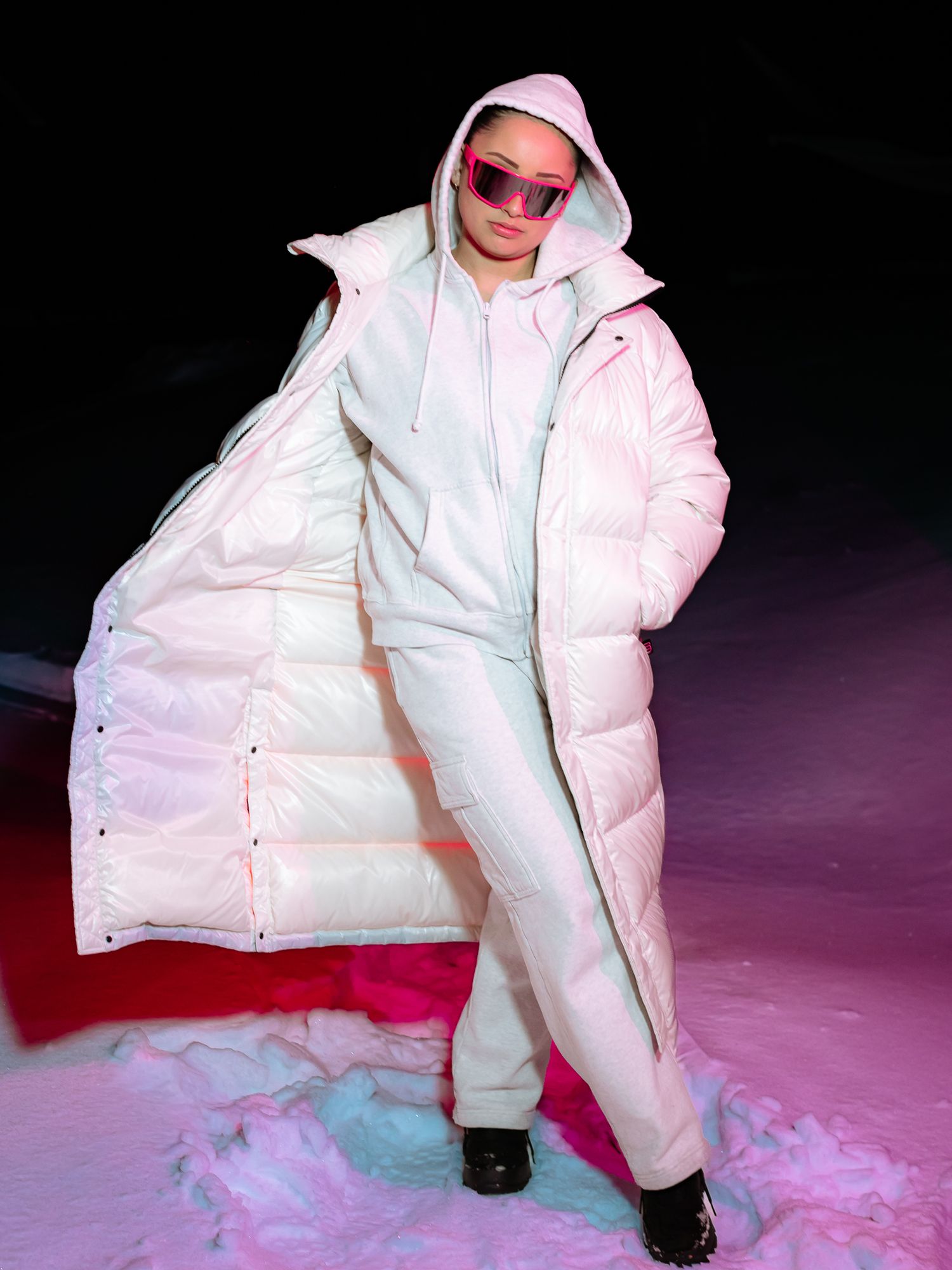 Woman in long white puffer coat and pink sunglasses posing in snowy outdoor setting at night.