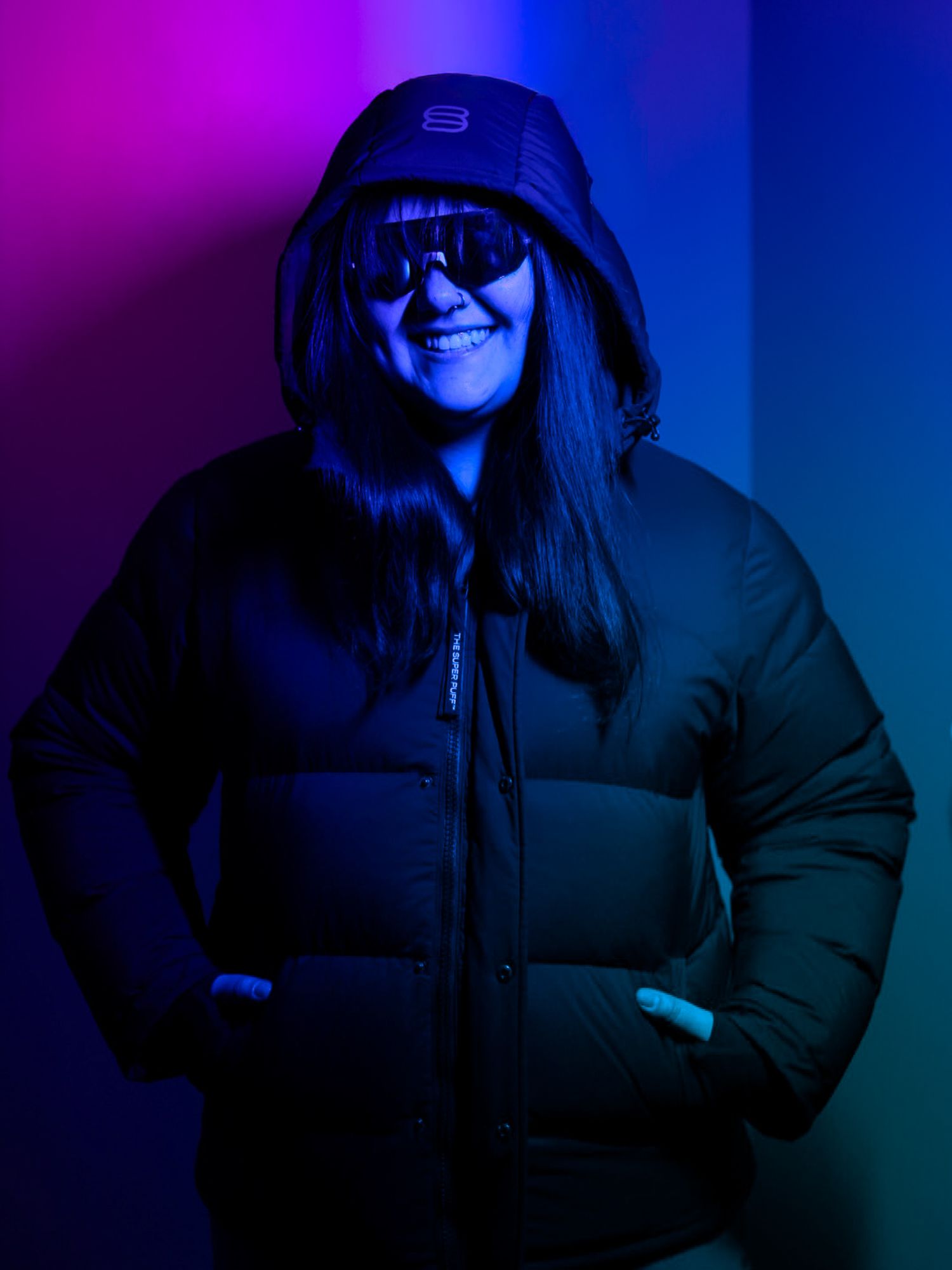 Woman in black hooded puffer jacket and shield sunglasses smiling under colorful neon lighting.