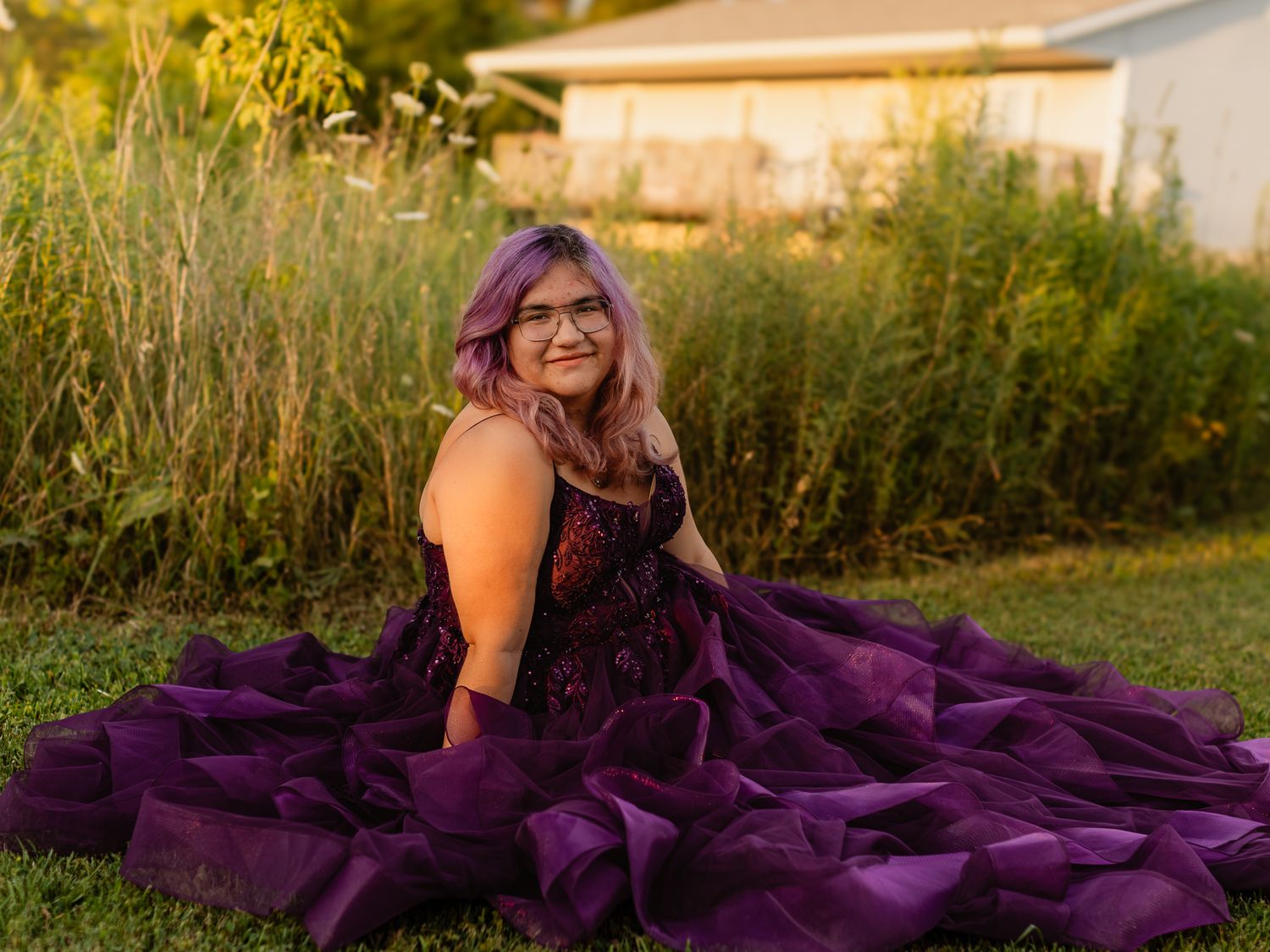 Young woman in stunning purple ball gown sitting on grass, featuring layered ruffles and floral details, with purple-tinted hair.