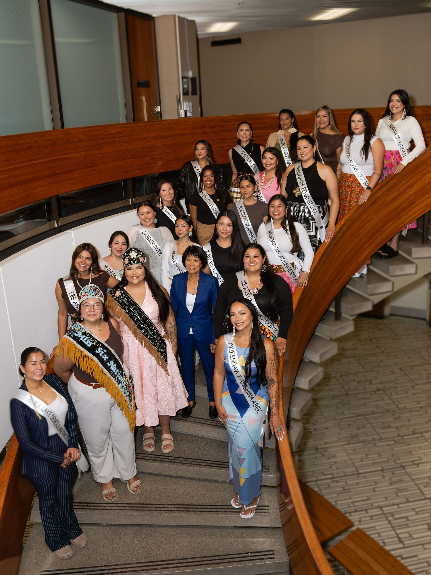 A large group of pageant contestants wearing sashes pose on a curved staircase in a formal building.