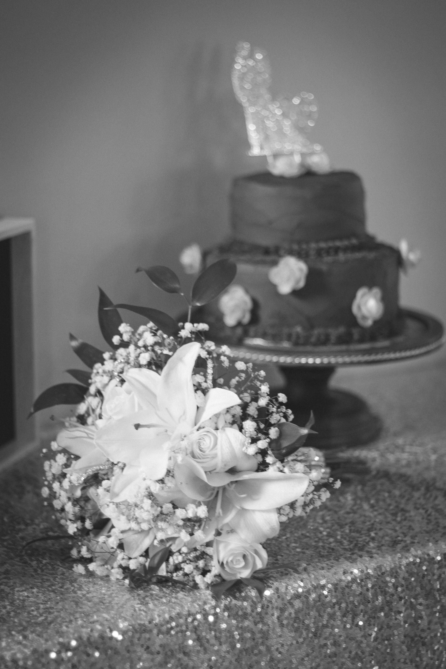 Black and white photo of bridal bouquet with roses and two-tier wedding cake in background.