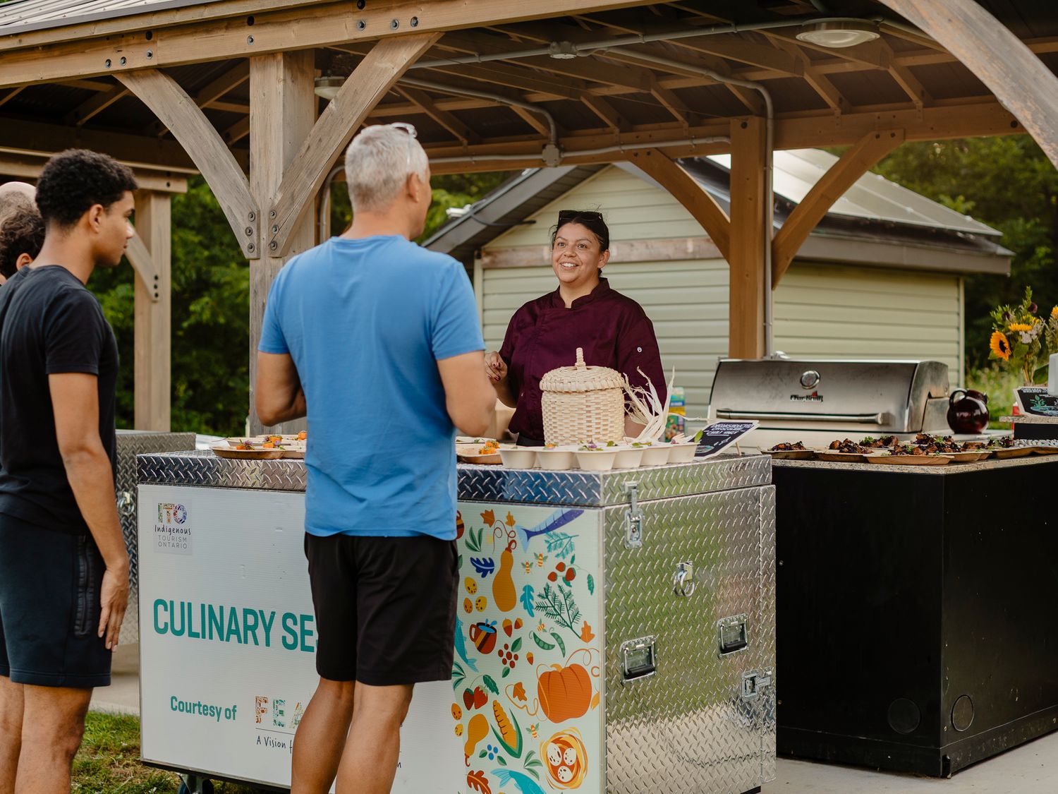 A chef serves guests at an outdoor culinary session booth under a wooden pavilion at a community event.