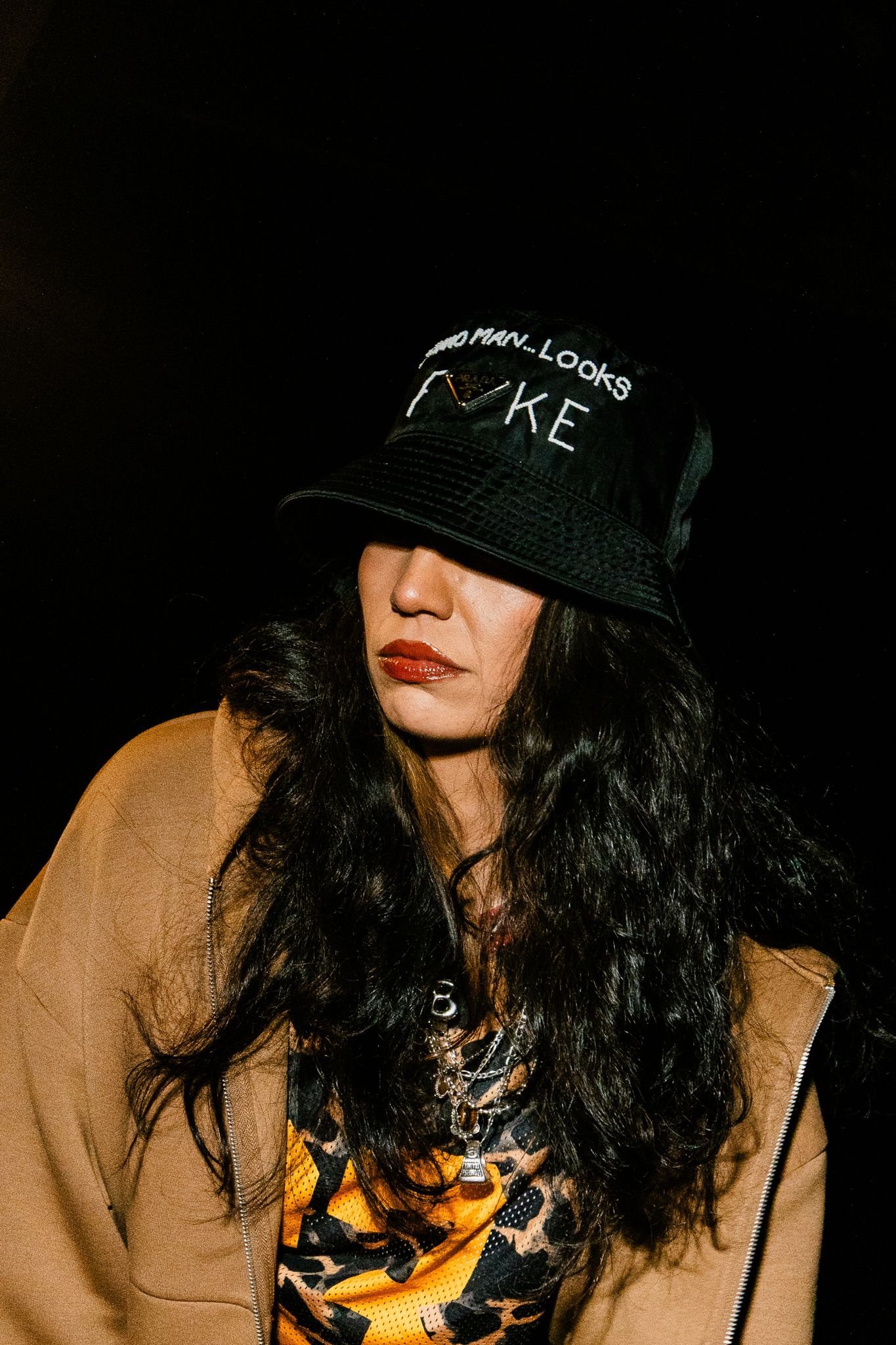 Woman in black bucket hat and camel coat with bold red lips against dark background.