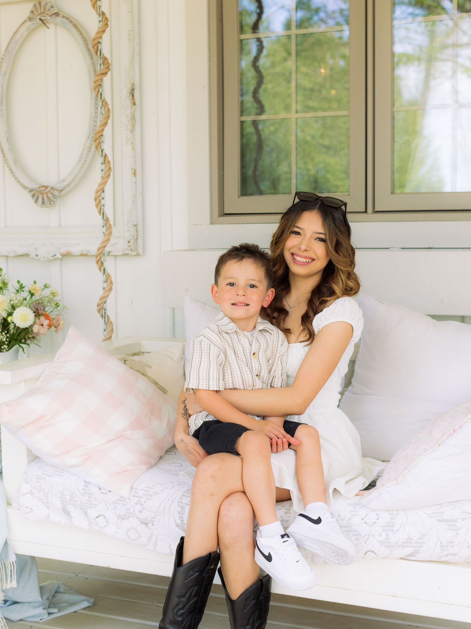 A smiling woman in white dress cuddles a young boy on an elegant white daybed on a charming porch.