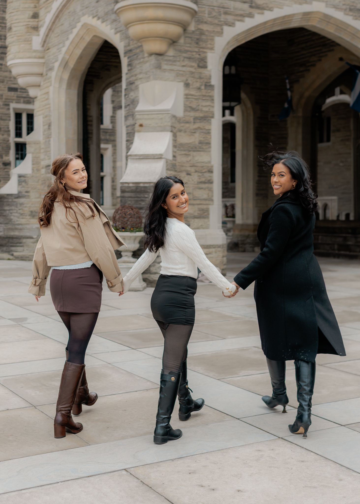 Three stylish women holding hands walk together in front of a stone building, wearing fall outfits with boots.