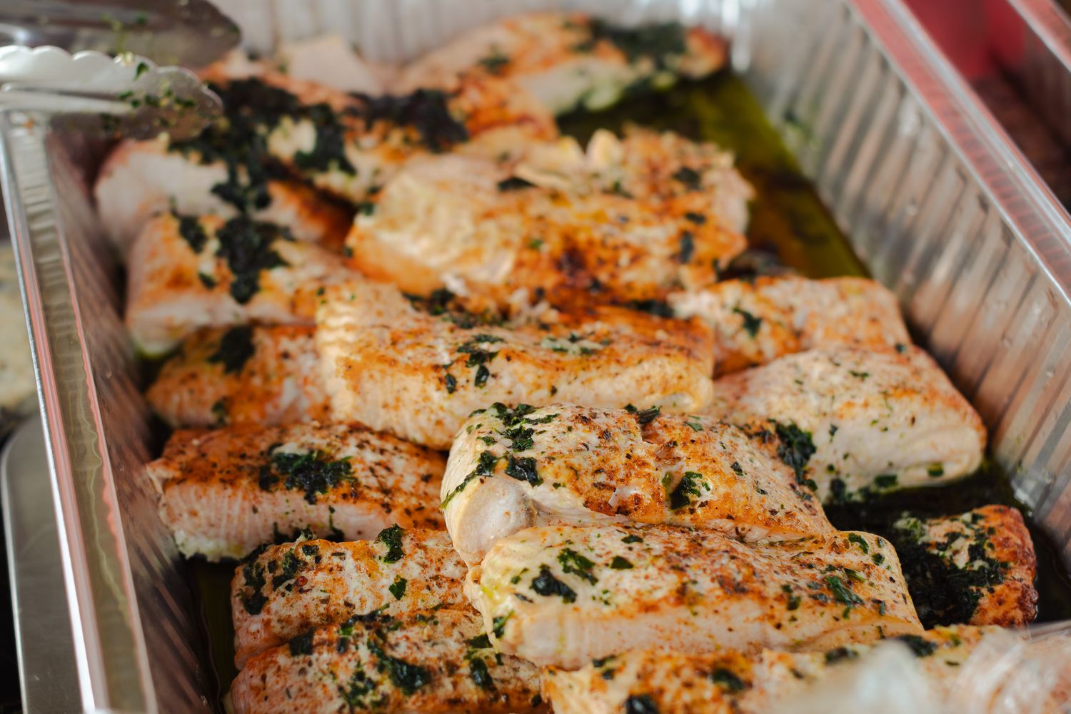 Seasoned baked fish fillets with herbs and spices served in a large aluminum catering tray.