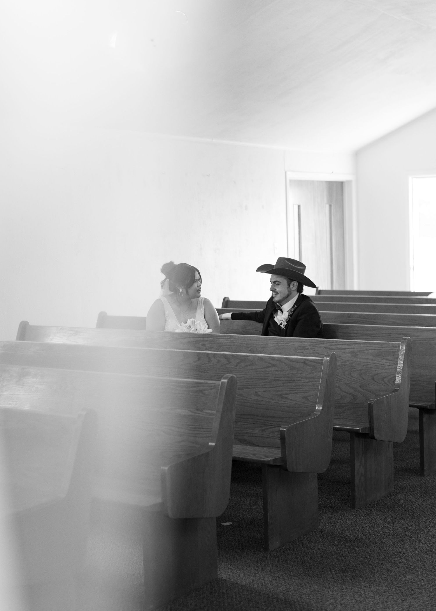 Black and white photo of a bride and groom in cowboy hat sitting together on church pews, sharing a quiet moment.