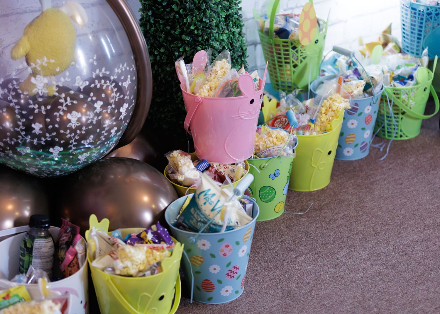 Colorful Easter buckets filled with treats and popcorn lined up on a table with festive balloons nearby.