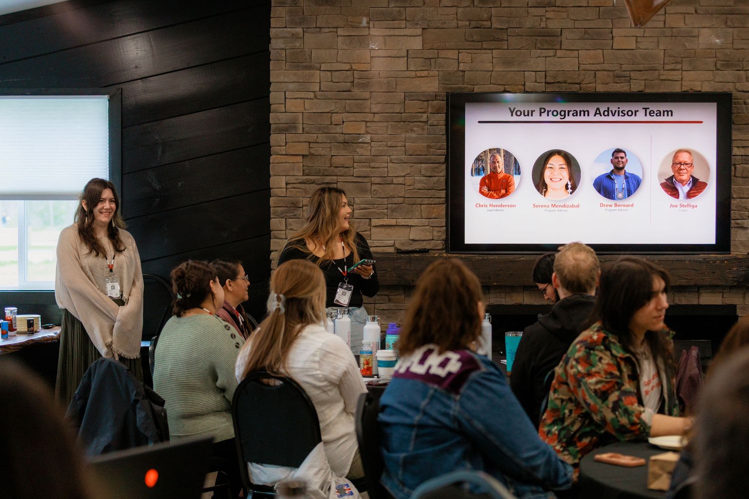 Two women present to a group at tables, with a Program Advisor Team slide displayed on a screen behind them.