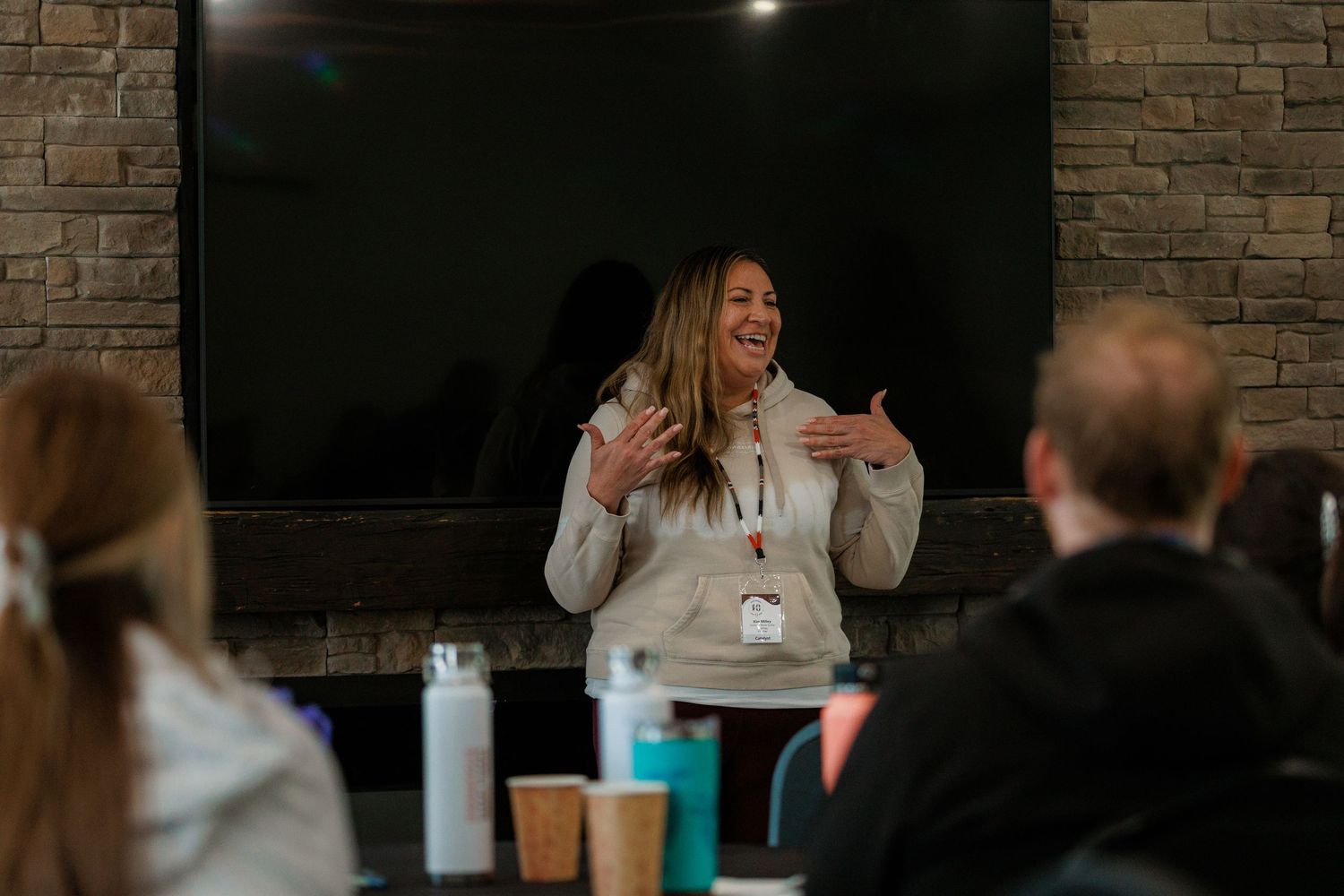 Woman presenting enthusiastically to seated audience in cozy stone-walled room with TV screen backdrop.