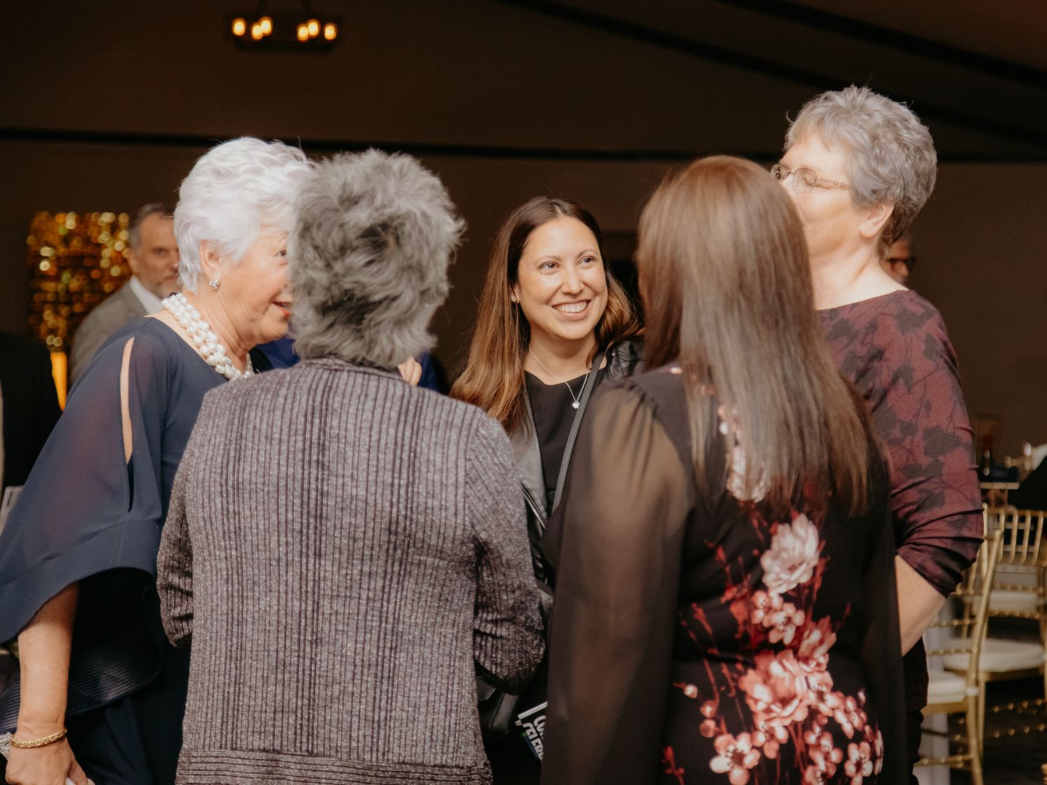 Group of elegantly dressed women socializing at a formal event in a warmly lit venue.