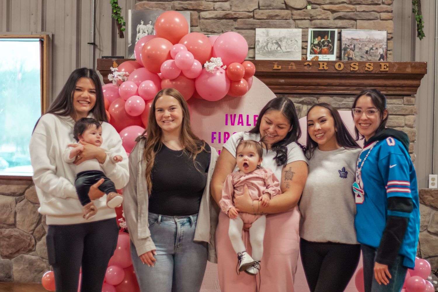 Group of women celebrating a first birthday party with pink balloons and festive decorations indoors.