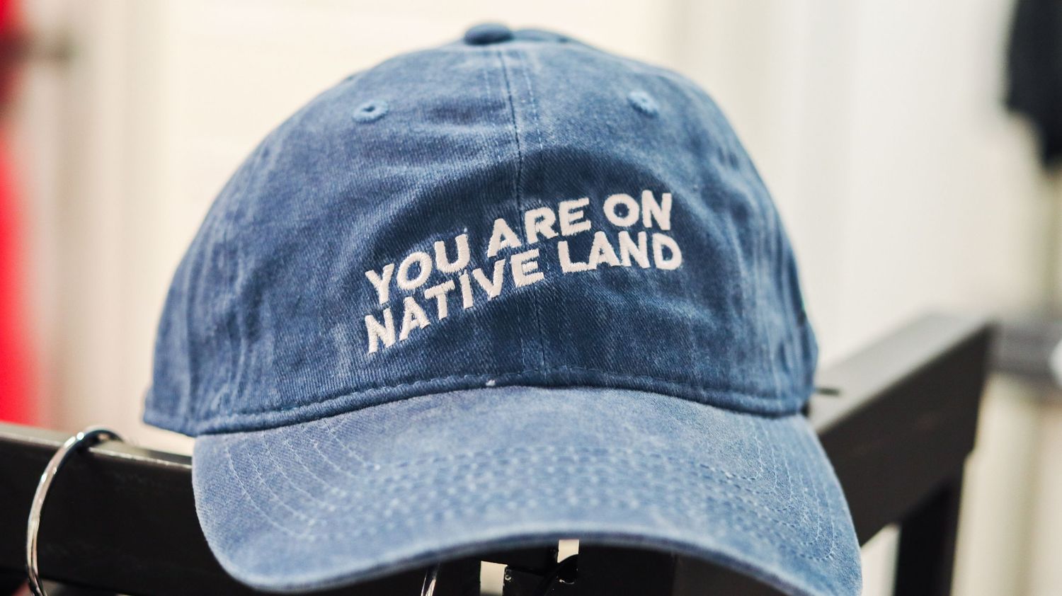Blue denim dad hat with white embroidered text reading YOU ARE ON NATIVE LAND displayed on a chair.