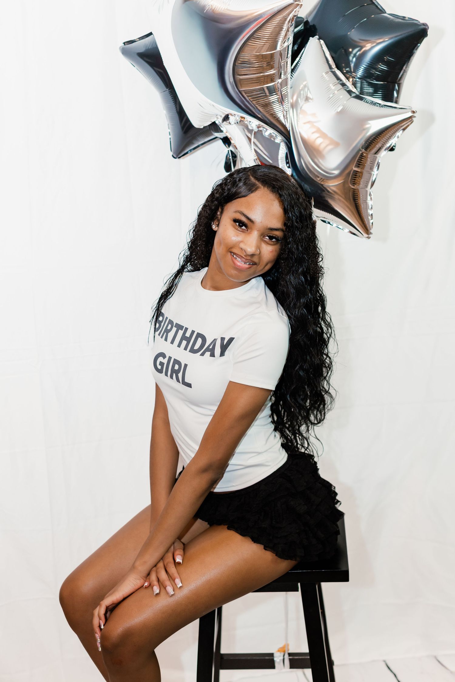 Young woman in a Birthday Girl t-shirt sits on a stool, smiling, with silver star balloons behind her.