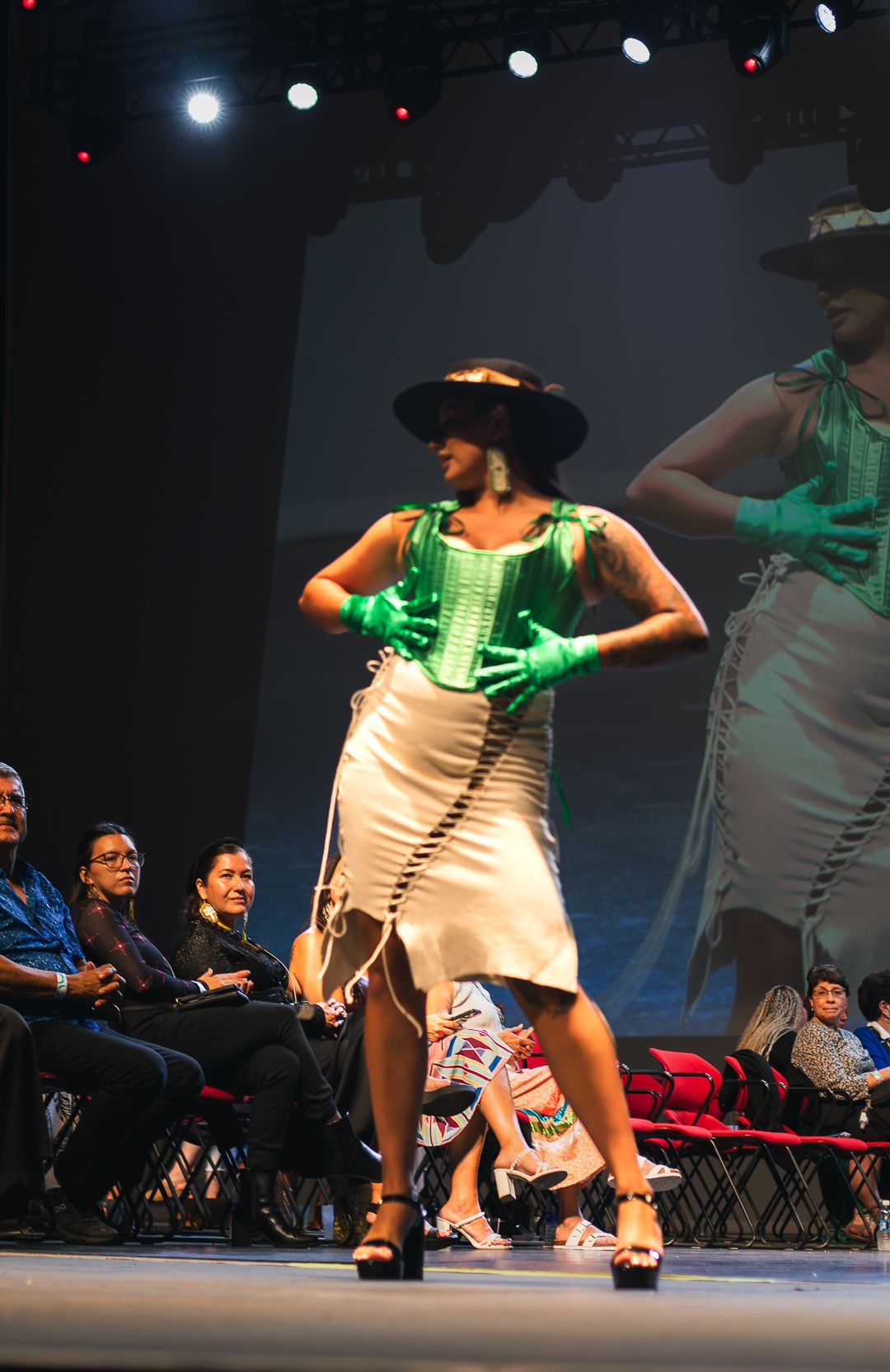 Model walks runway in white skirt, green corset, black hat, and gloves at a fashion show with seated audience.
