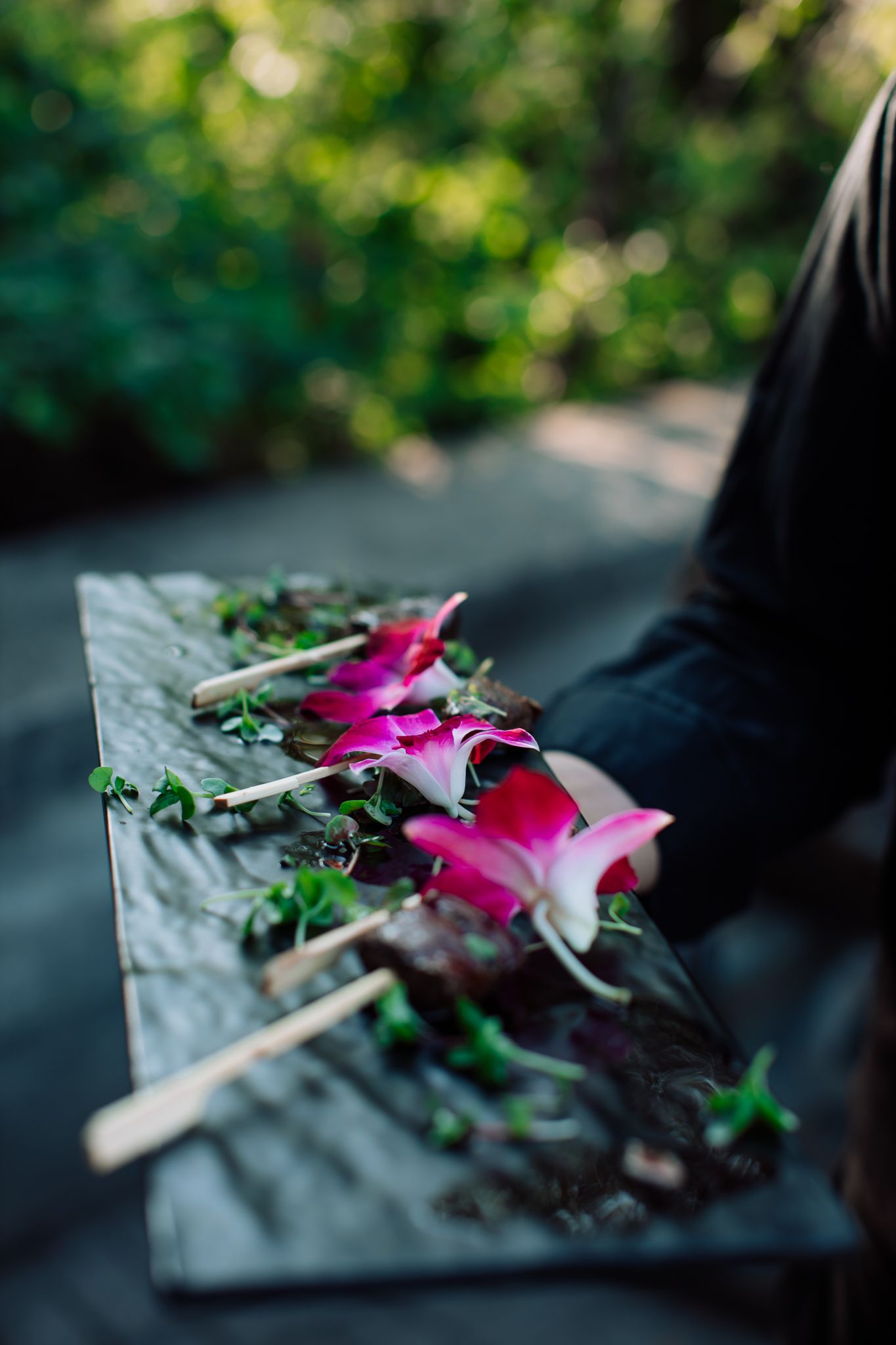 Pink rose petals and green herbs arranged on wooden skewers on a rustic gray board outdoors.