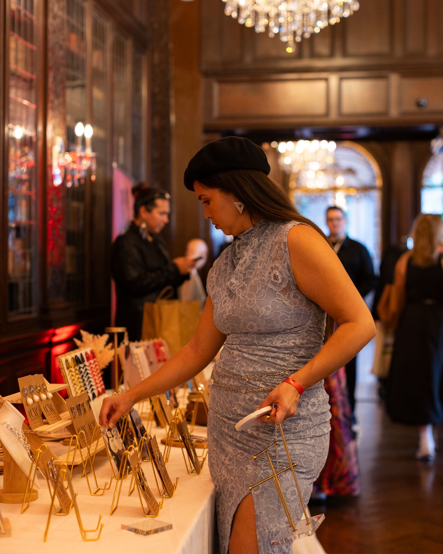 Elegant woman in silver beaded gown browsing gold trophy displays in an ornate chandelier-lit ballroom.