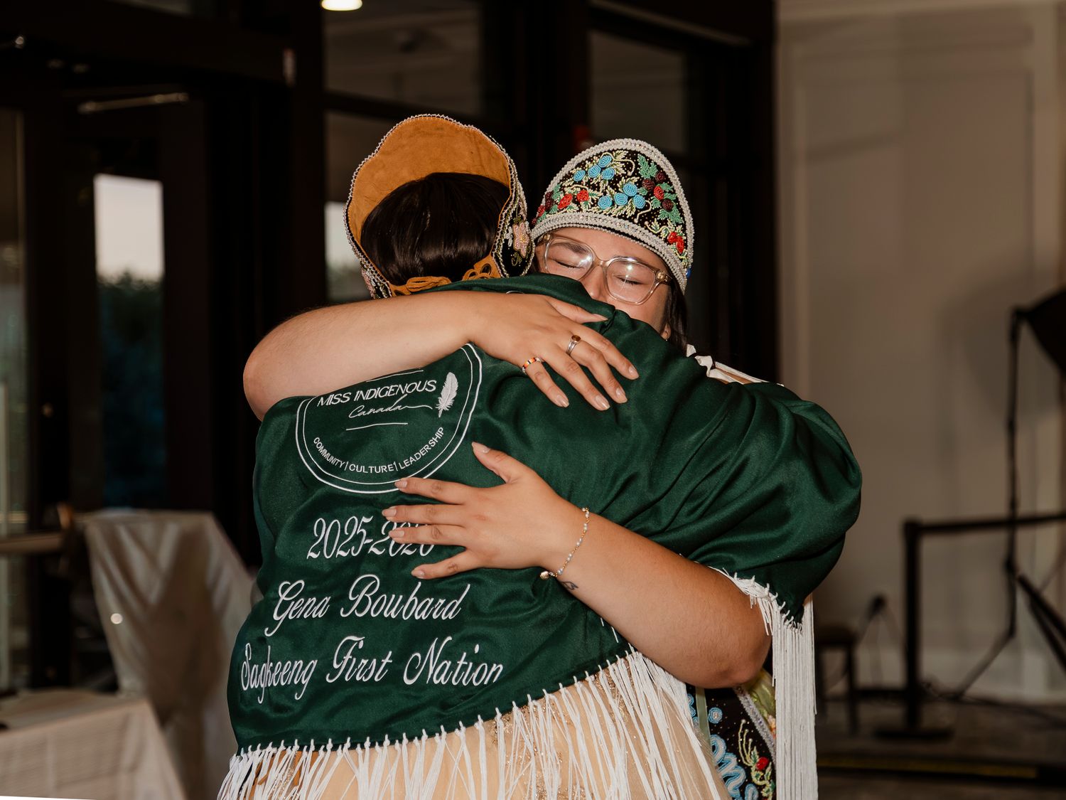 Two people share a warm embrace, one wearing a green jacket reading Nuchang First Nation 2025.