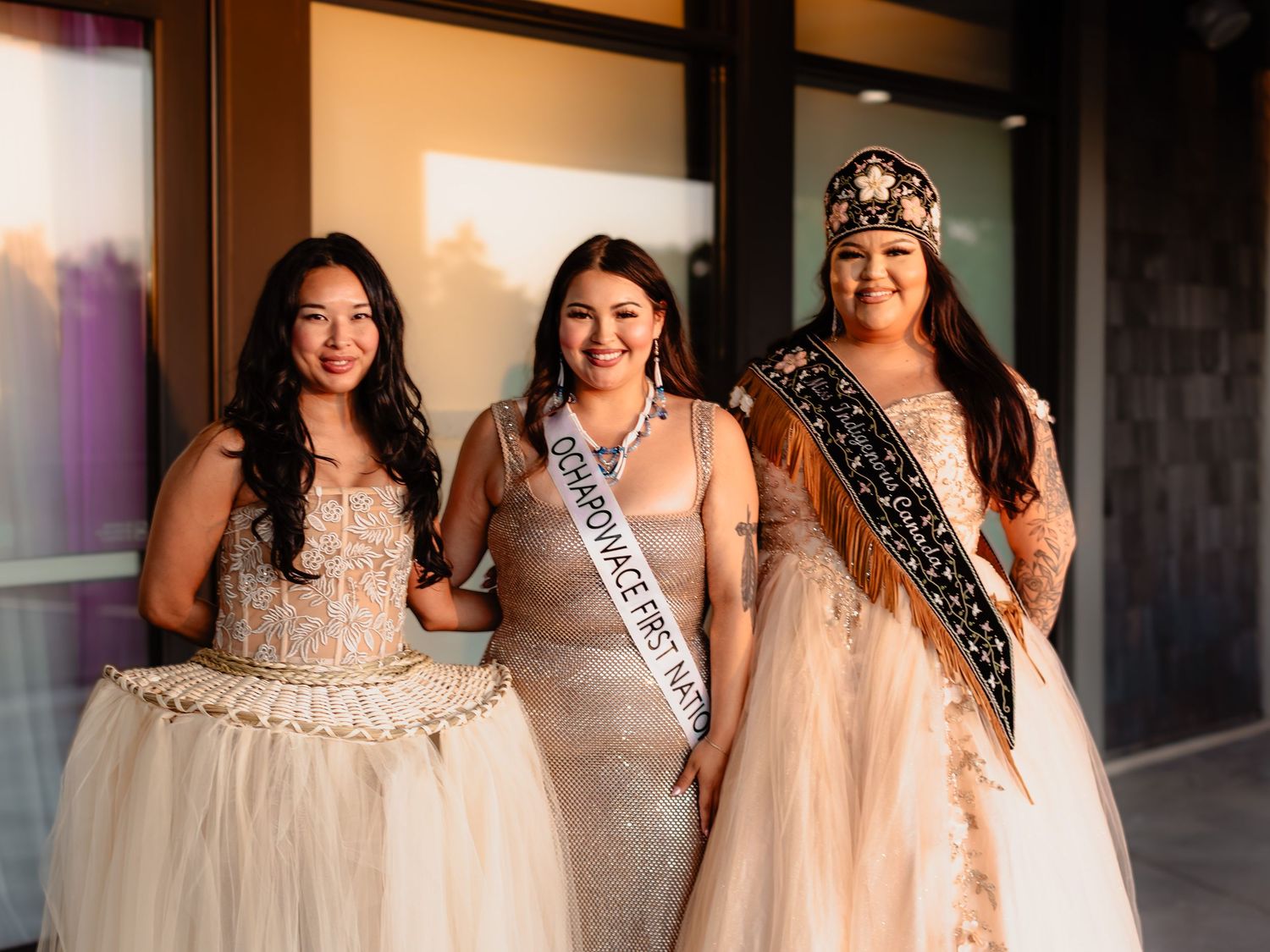 Three women in elegant gowns pose together, one wearing a Miss Indigenous First Nations sash and crown.