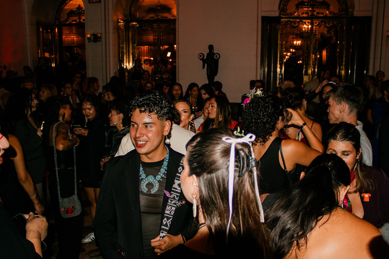 Young man smiling at a lively indoor evening party surrounded by guests in a warmly lit ornate venue.