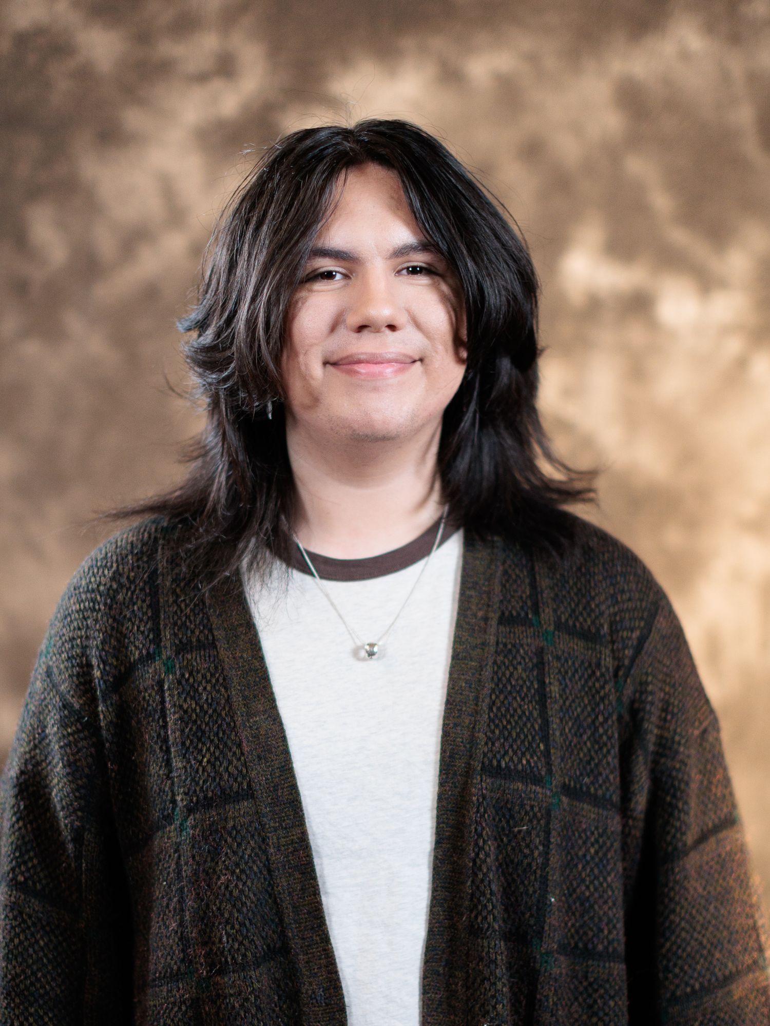 Young person with long dark hair wearing a green plaid cardigan and necklace smiling against a brown backdrop.