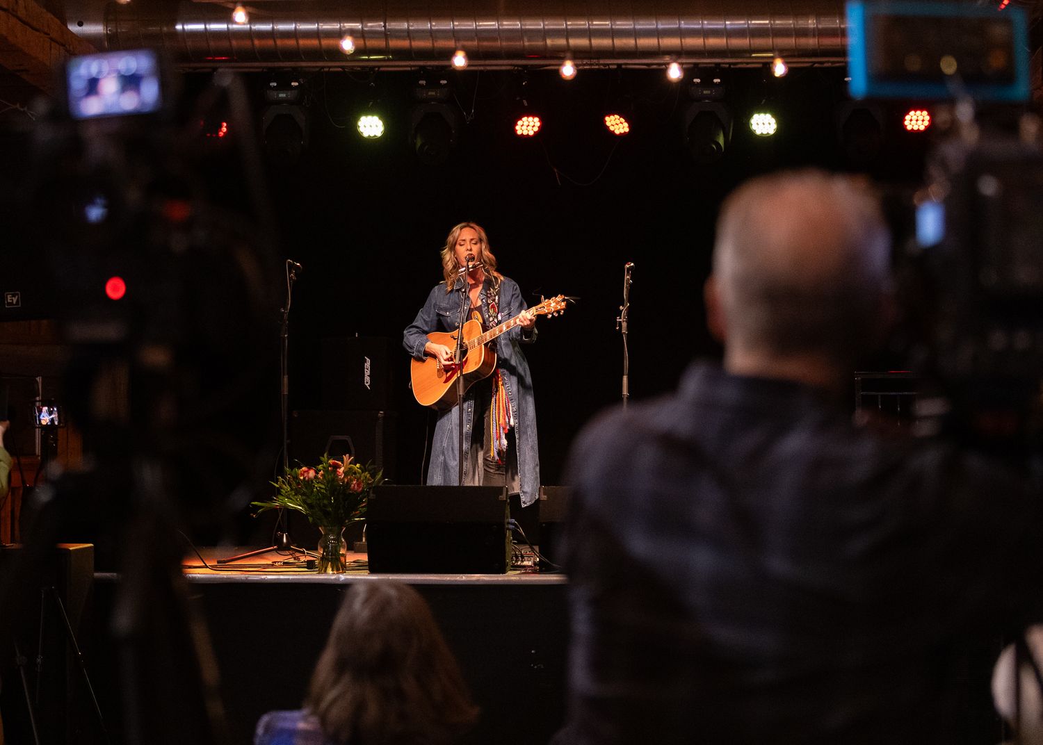 A female musician plays acoustic guitar and sings on a lit stage while being filmed by a videographer in the foreground.