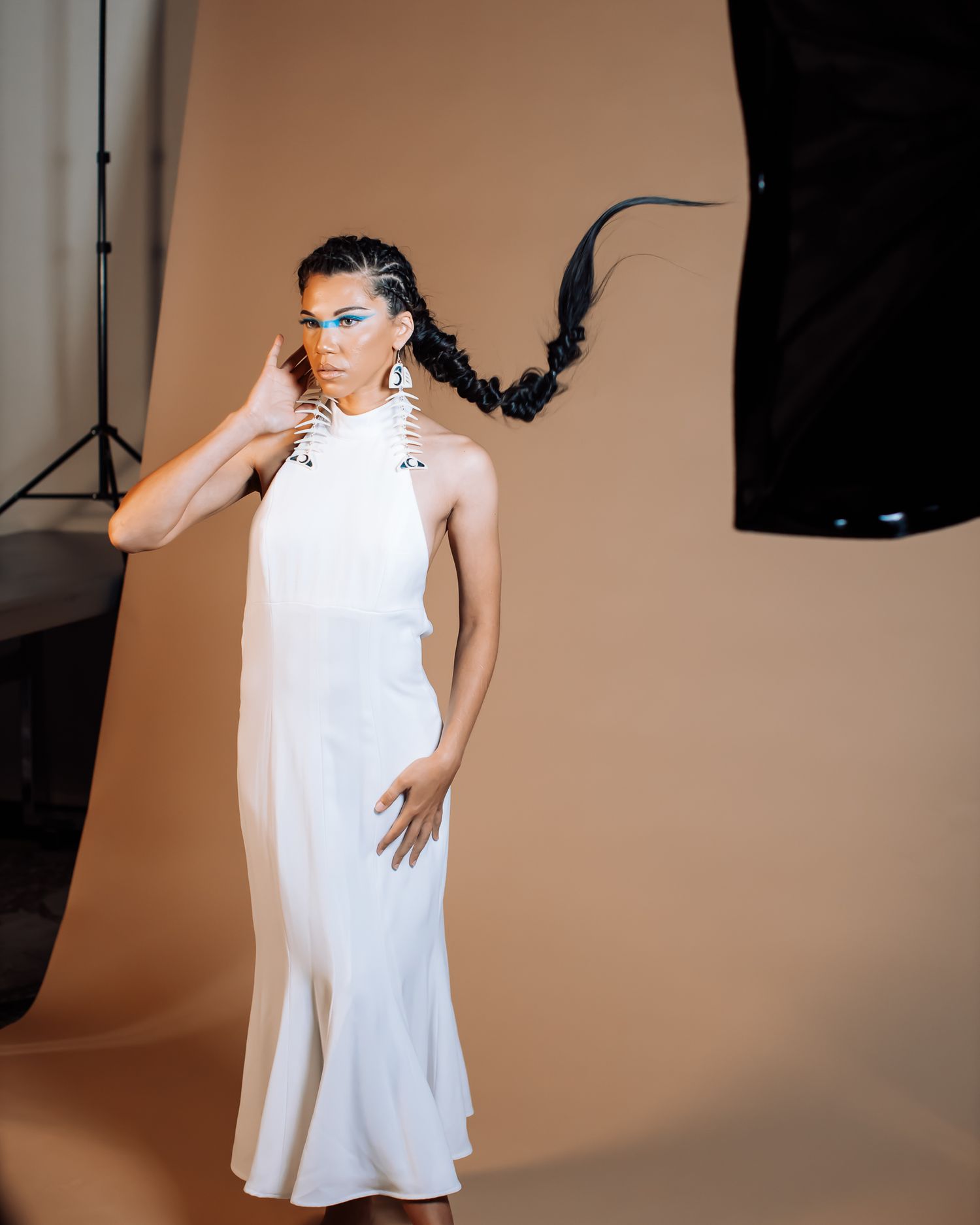 Woman in white halter maxi dress with dramatic braided updo and bold eye makeup in photo studio.