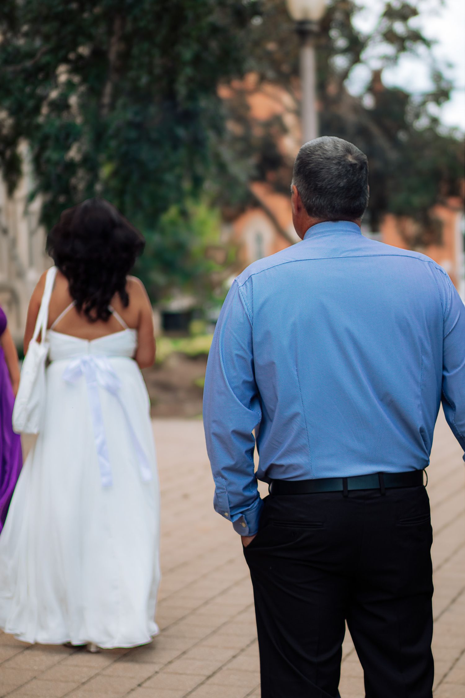 Groom in blue shirt walks behind bride in white wedding gown on outdoor brick pathway.
