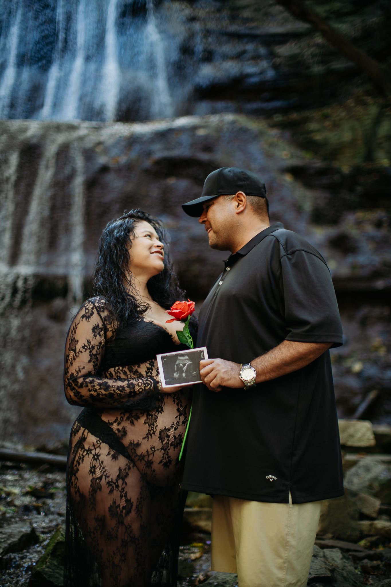 A couple shares an intimate moment by a waterfall as they hold a red rose together.