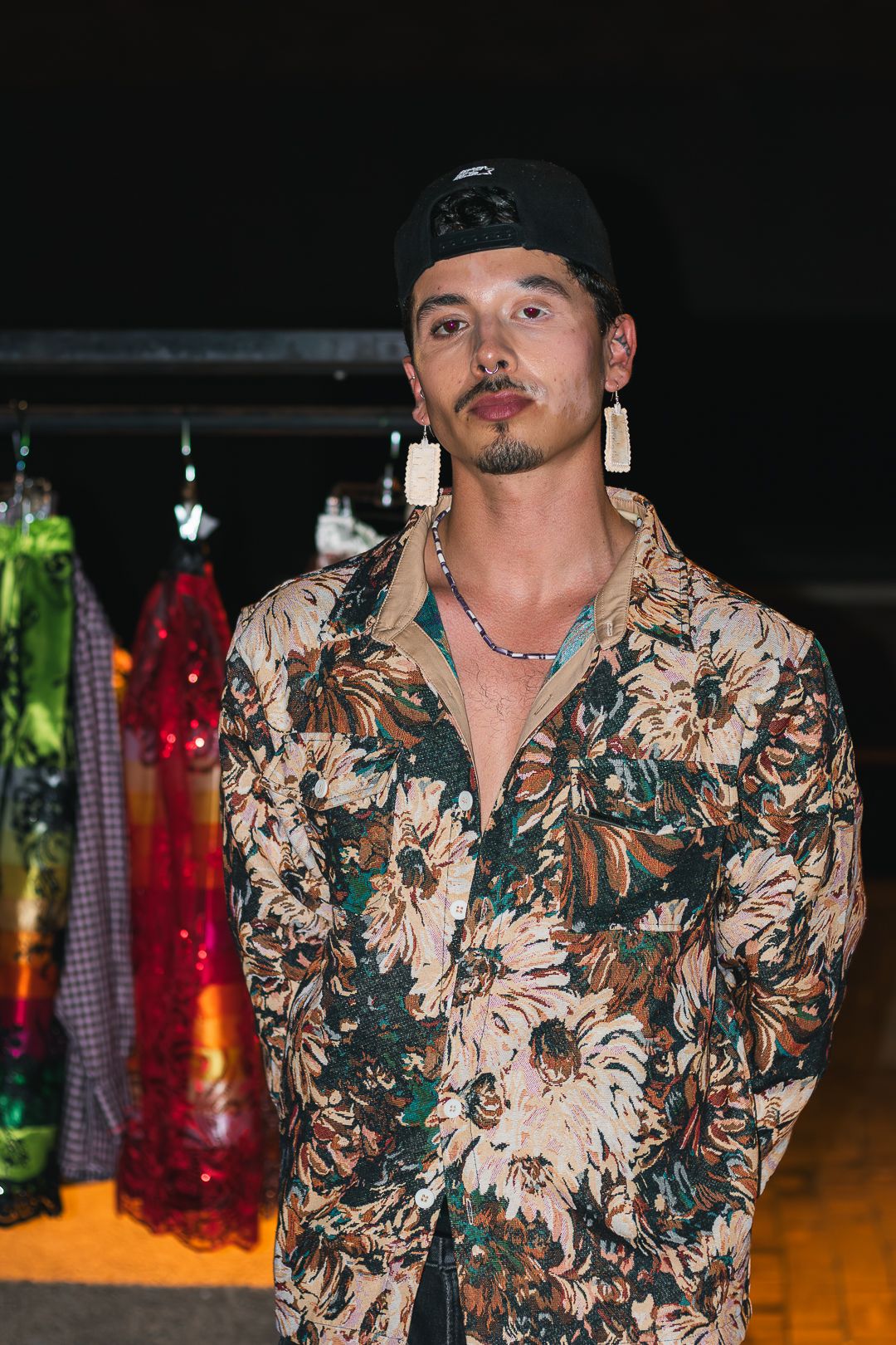 Person wearing a floral print shirt poses in a dark indoor setting with colorful lights in the background.