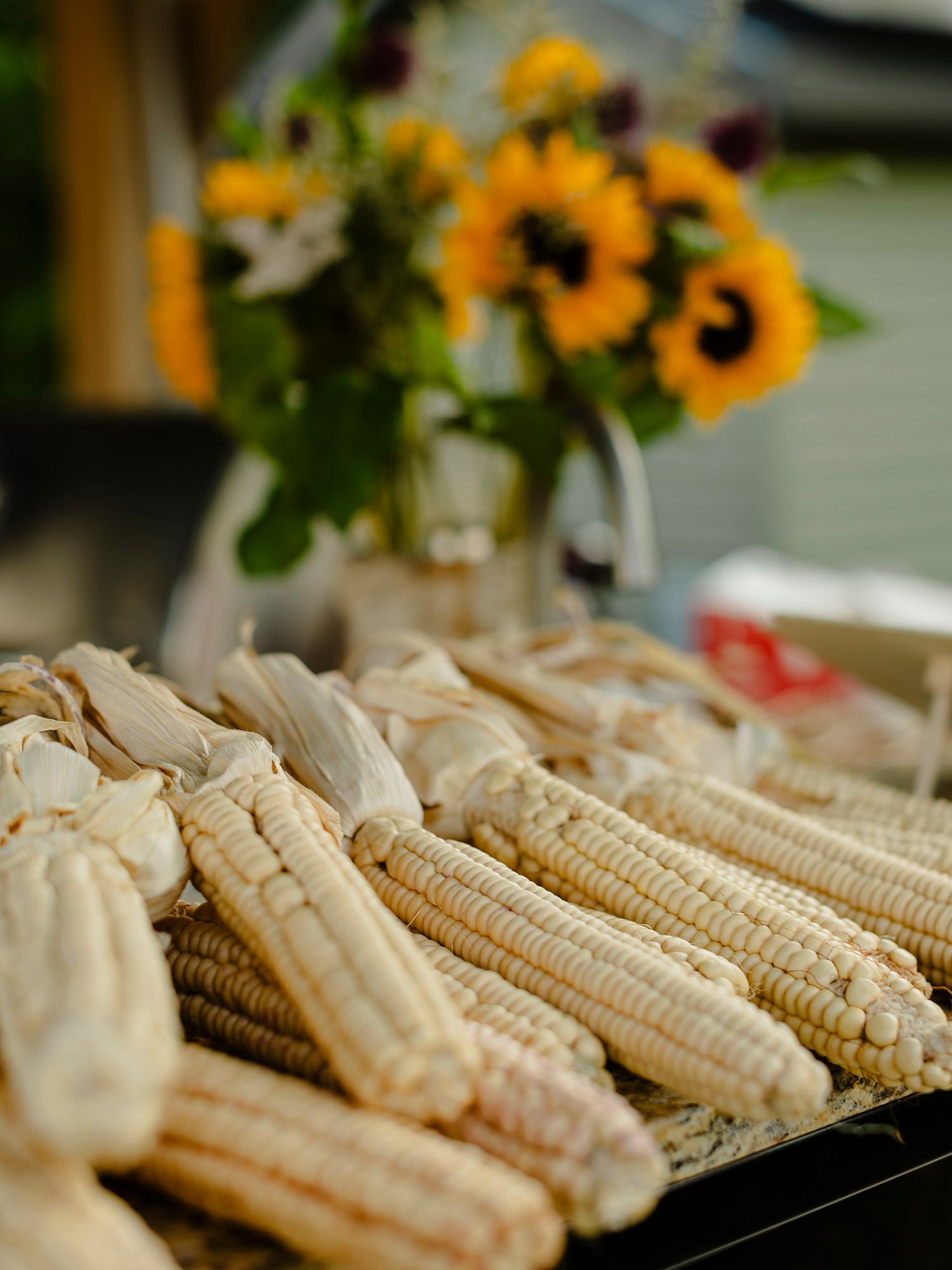 White corn on the cob displayed at a farmers market with sunflowers in a vase in the background.