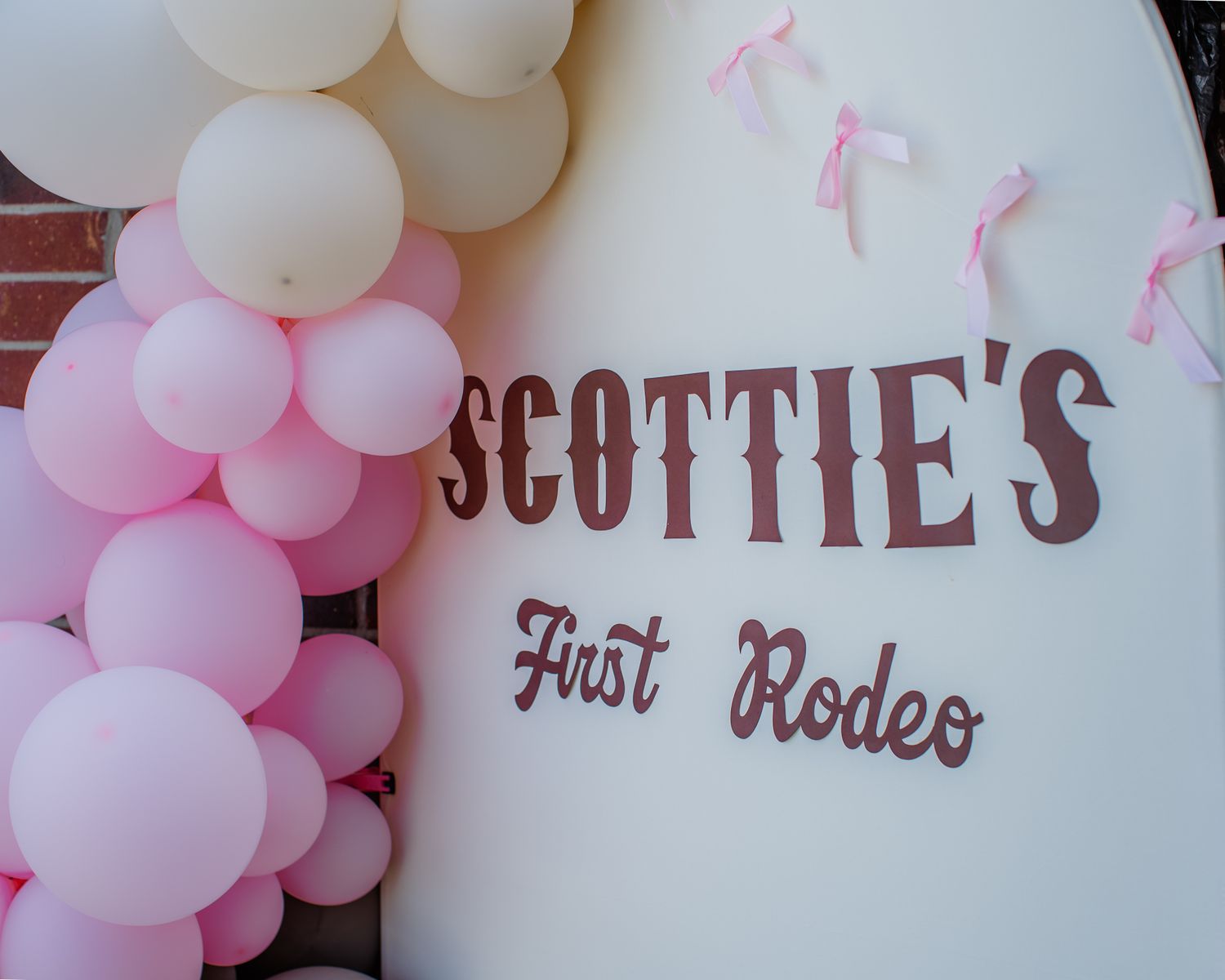 Pink and white balloon arch decorates a sign reading Scottie's First Rodeo at a baby's first birthday party.