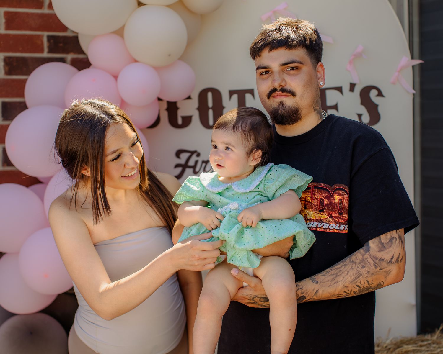 Young couple smiling and holding their baby in green outfit in front of pink balloon first birthday backdrop.