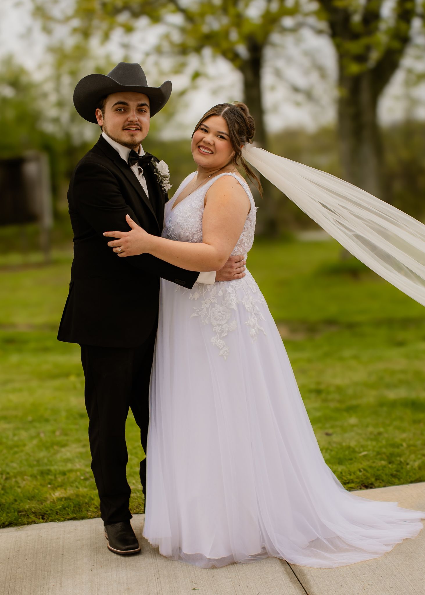 Bride in white lace gown with flowing veil embraces groom wearing black cowboy hat and suit outdoors.