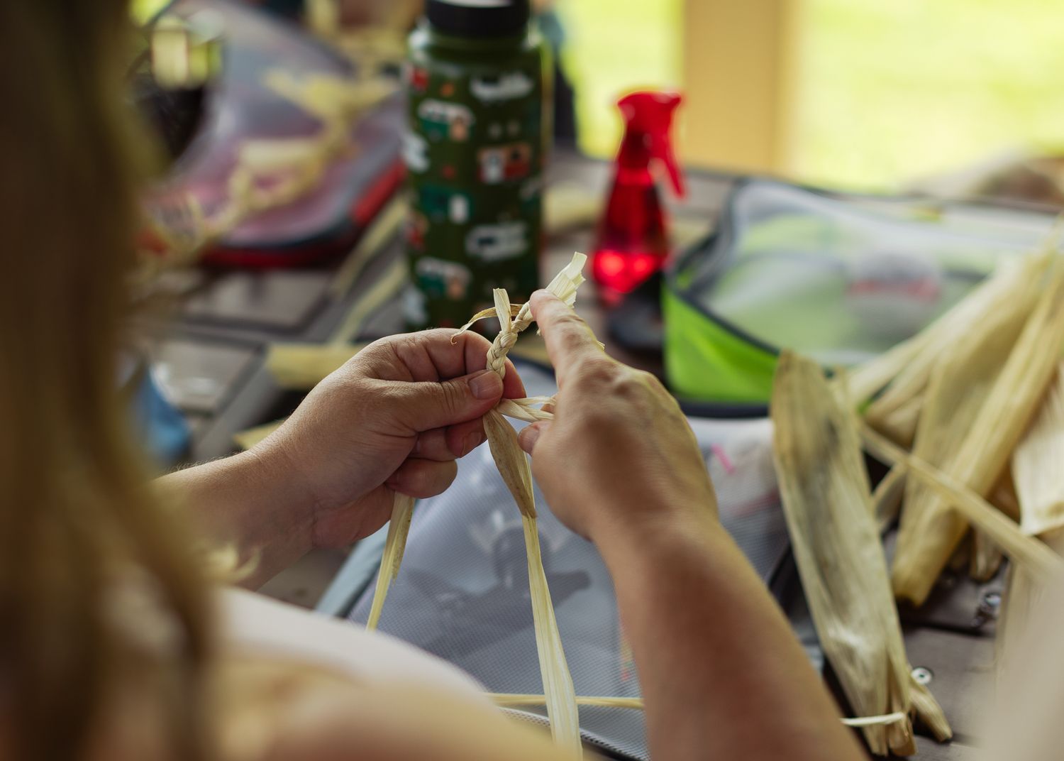 Close-up of hands weaving natural fibers at a craft table with supplies in background.