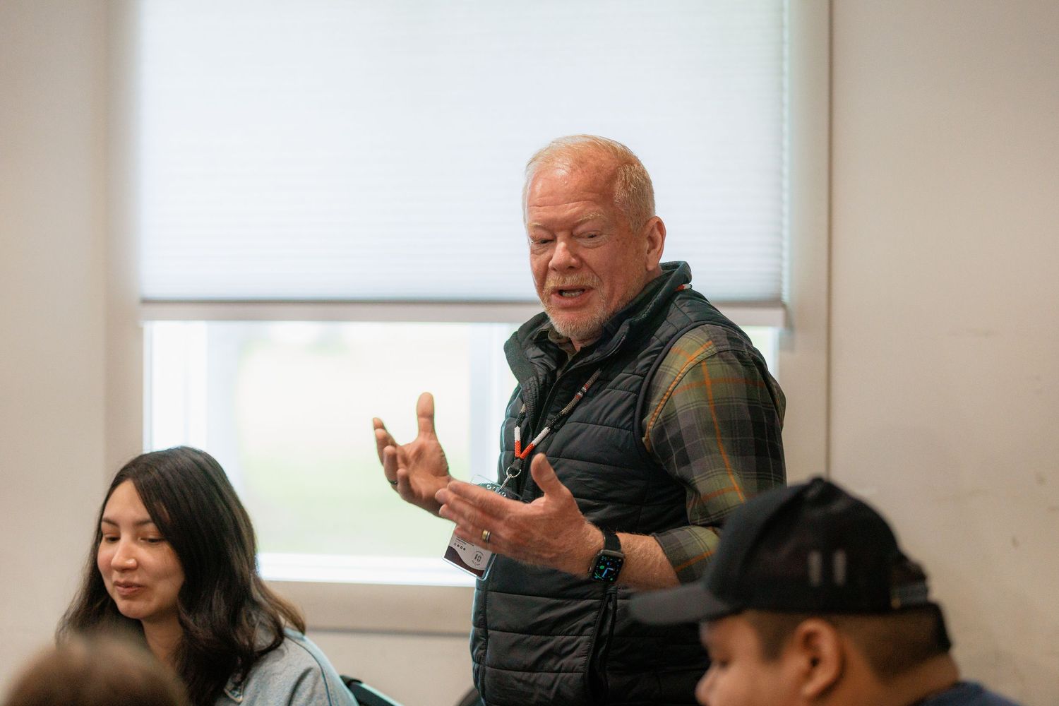 Older man gesturing while speaking to a group of students in a classroom setting.