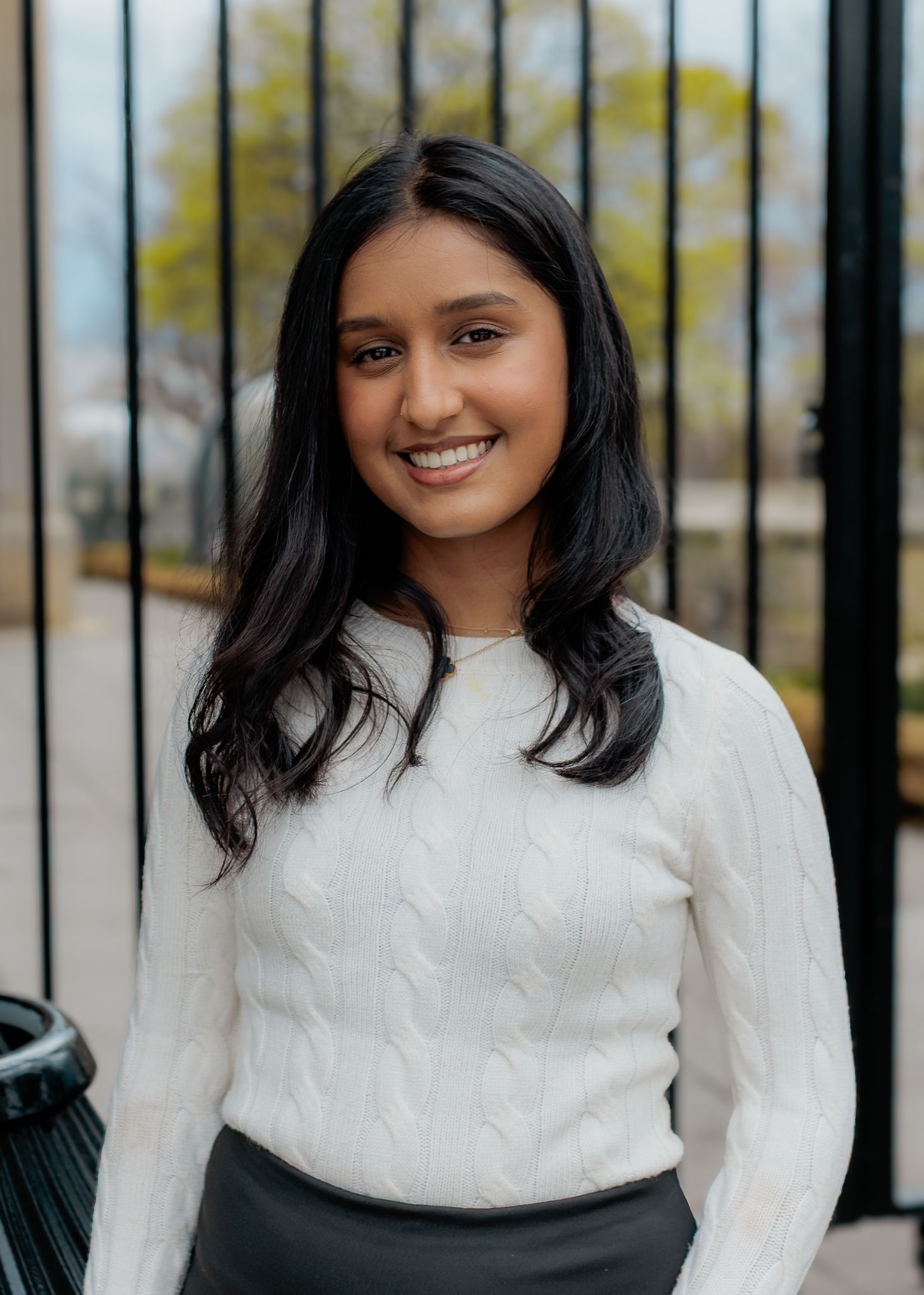 Young woman in white cable-knit sweater smiling outdoors near black iron gate with trees in background.