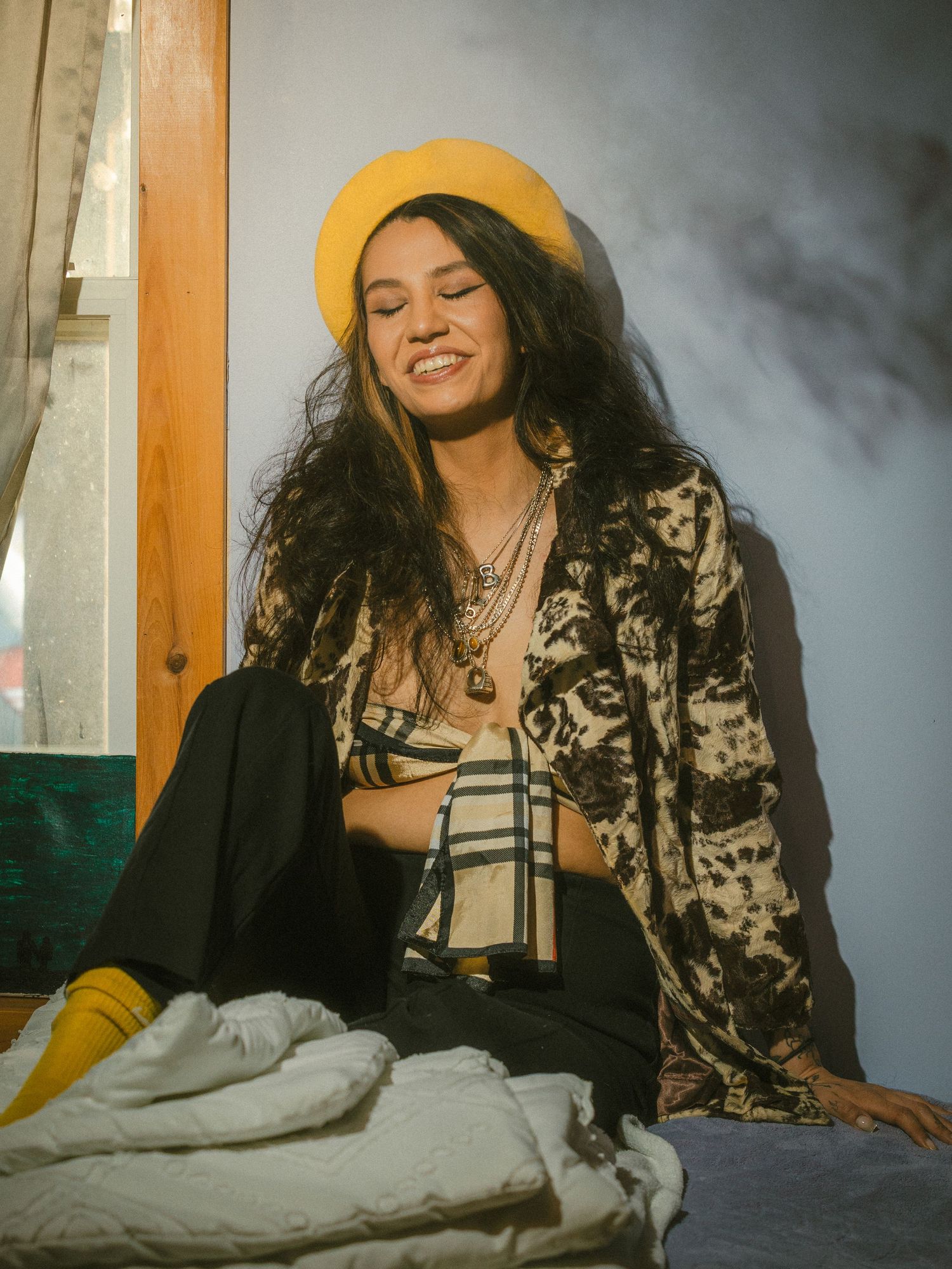 Smiling woman in yellow beret and floral jacket sitting in sunlit room.