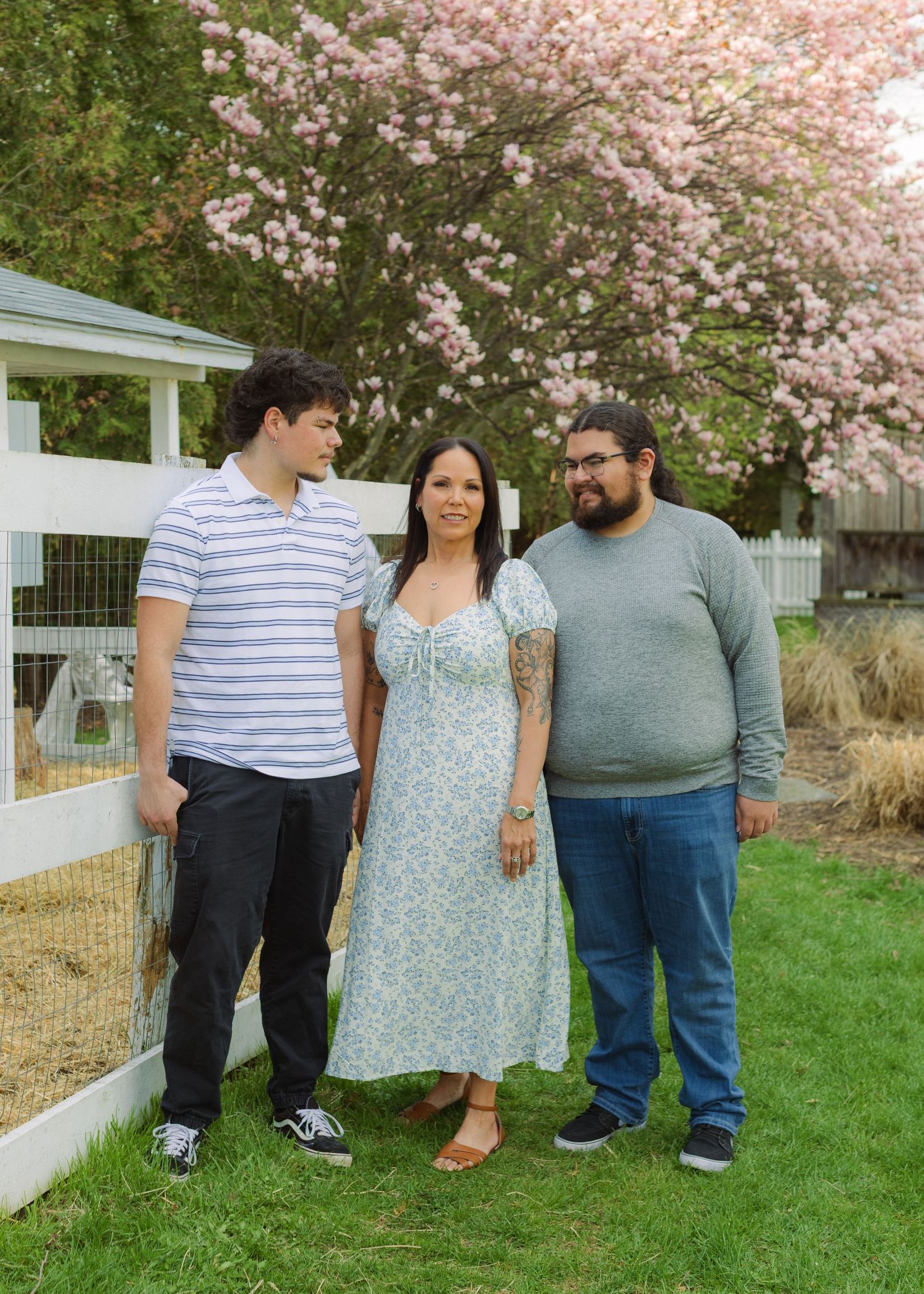 Three people smiling outdoors near a white fence with pink cherry blossom tree in background.