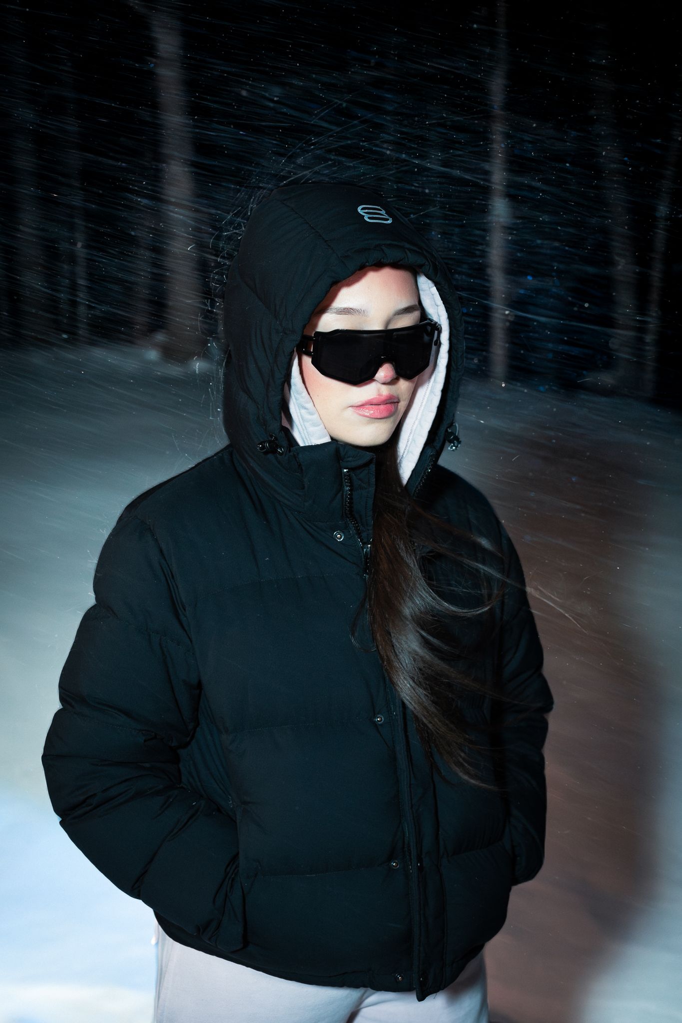 Woman in black hooded puffer jacket and dark sunglasses standing in snowy forest at night.