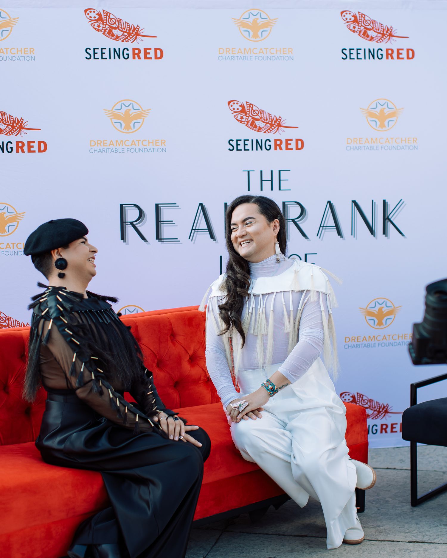 Two women in contrasting black and white outfits chat on a red sofa at The Real Frank House event.