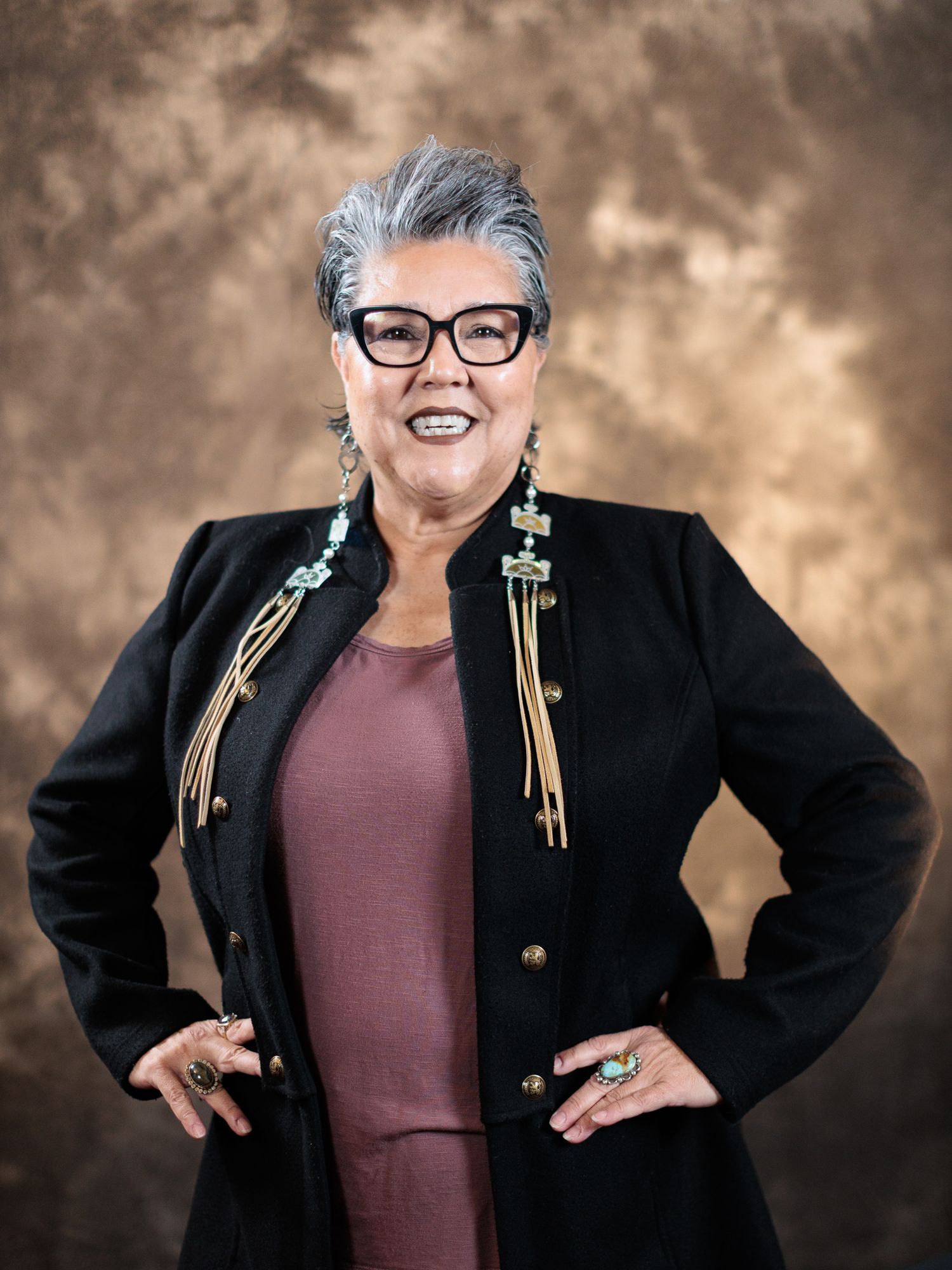 Confident older woman with short gray hair wearing a black embroidered jacket and turquoise jewelry poses with hands on hips.