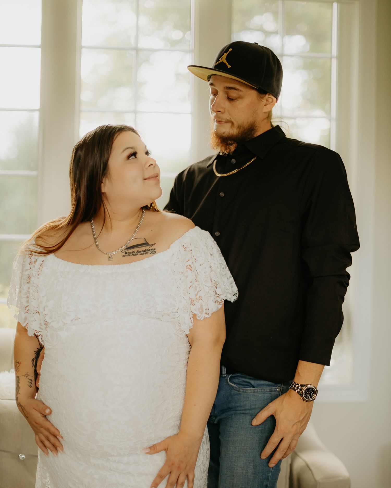 Couple in casual attire poses together in bright sunlit room with white decor.
