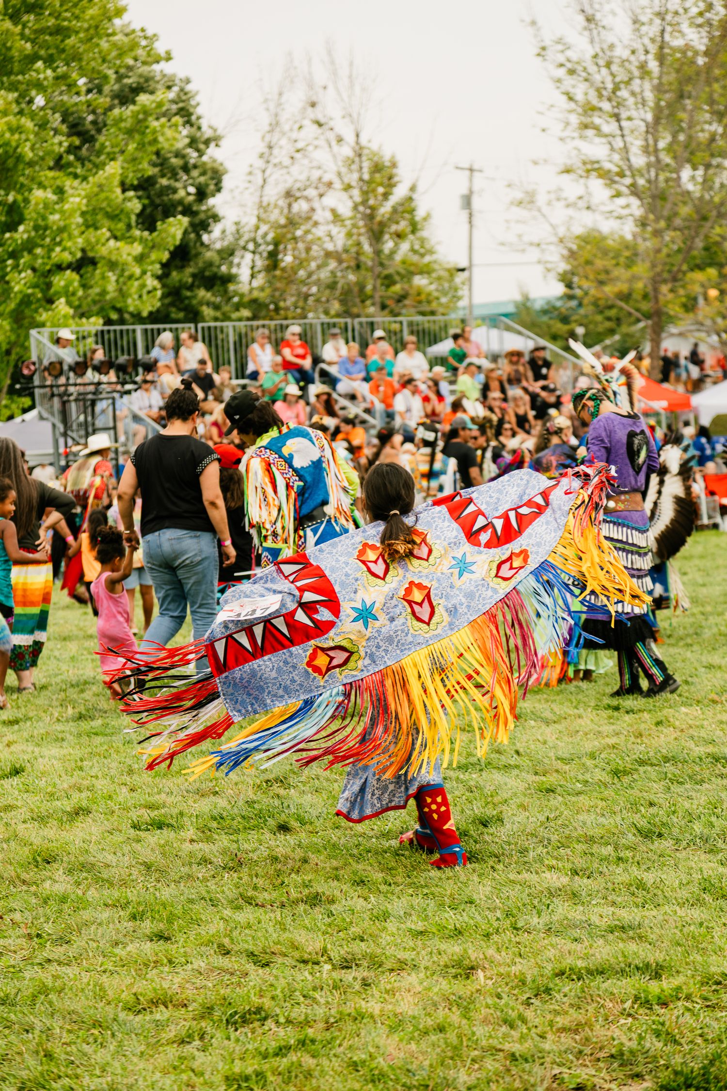 Vibrant traditional dancers in regalia perform during a powwow on a grassy field with spectators watching.