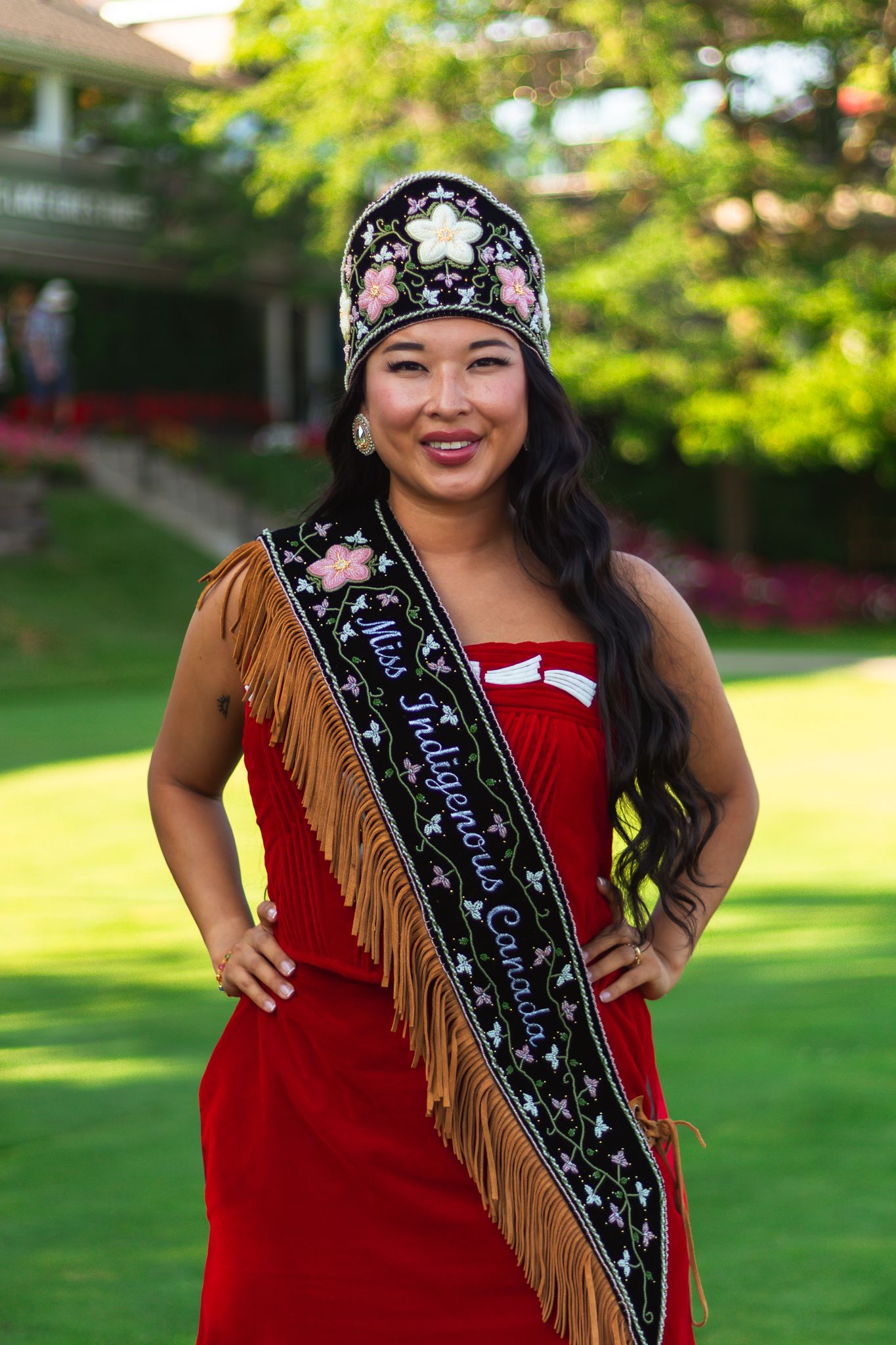 Person in traditional regalia with decorative sash outdoors on green lawn.