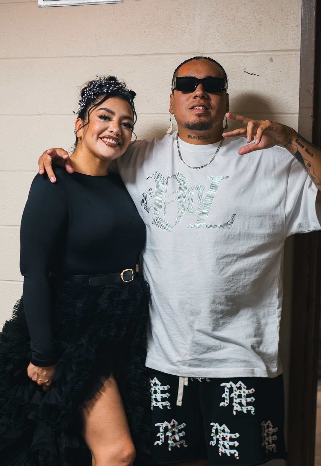 Two people posing together in contrasting black and white outfits against a light colored wall.