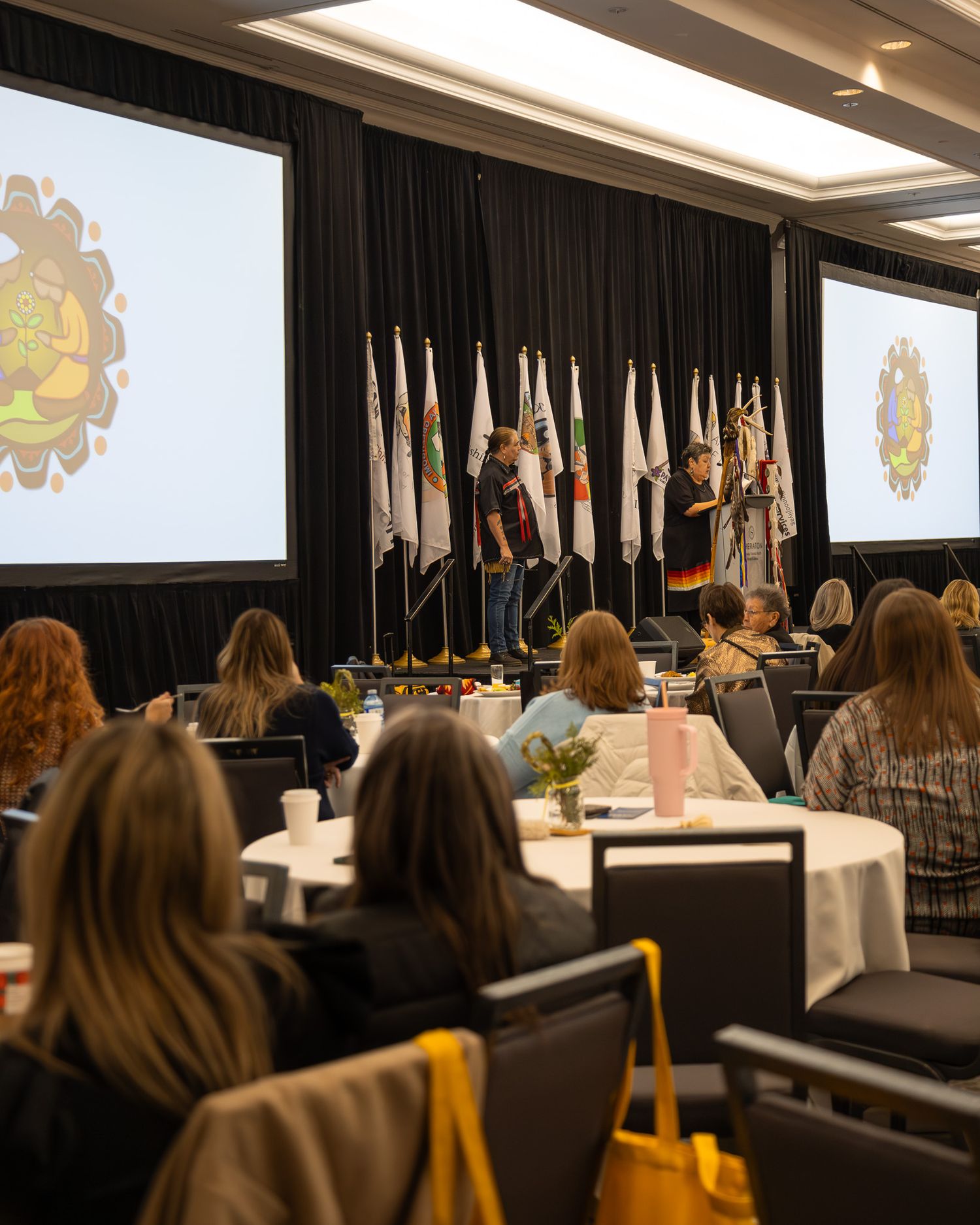 Conference room set up with round tables and chairs facing a stage with flags and decorative backdrop.
