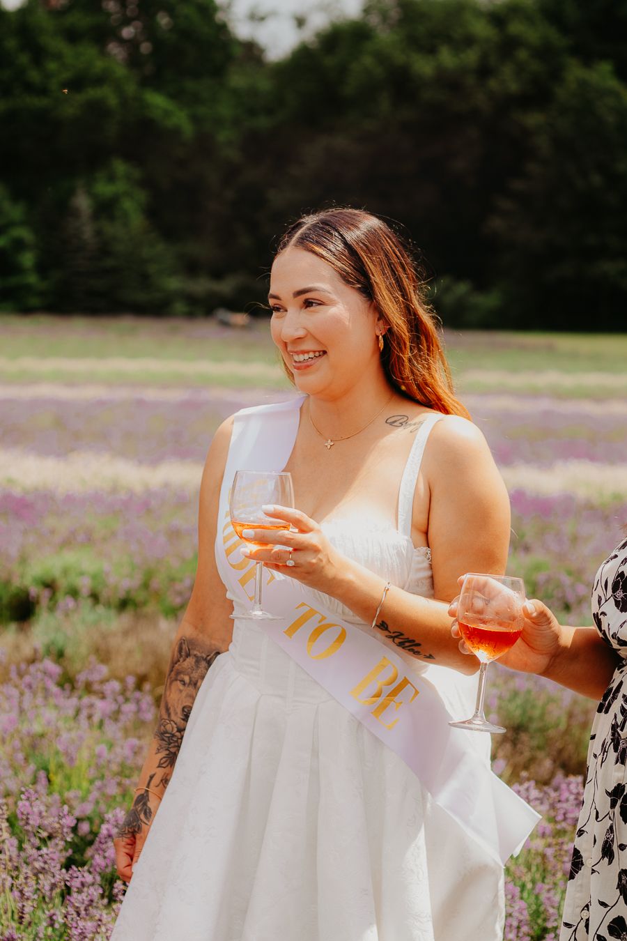 Woman in white dress holding wine glass smiling in lavender field at sunset.